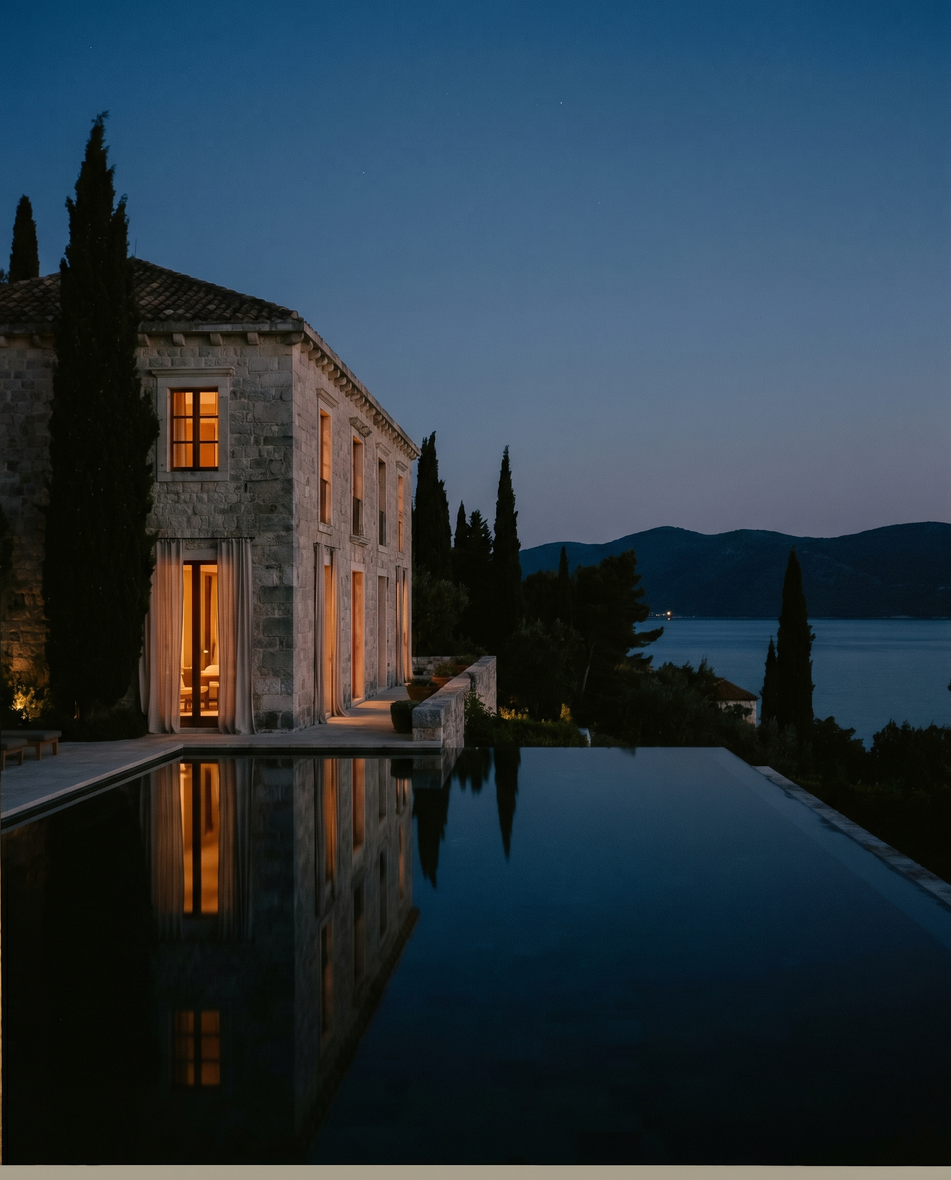 An Aman-style limestone villa at blue-hour twilight with a still reflecting pool and warm lamplight.