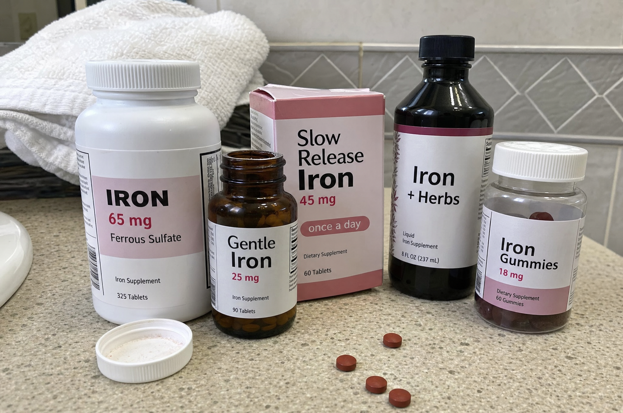 A lineup of common over-the-counter iron supplements crowded on a bathroom counter: ferrous sulfate tablets, gentle iron, a slow-release iron box, liquid iron with herbs, and iron gummies, with a few loose tablets spilled beside them