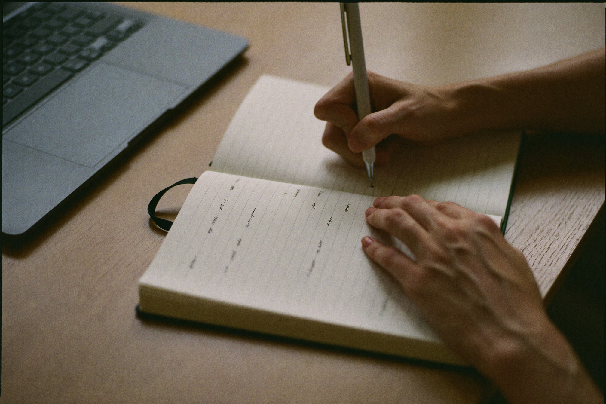 Hands writing in a notebook beside a closed laptop