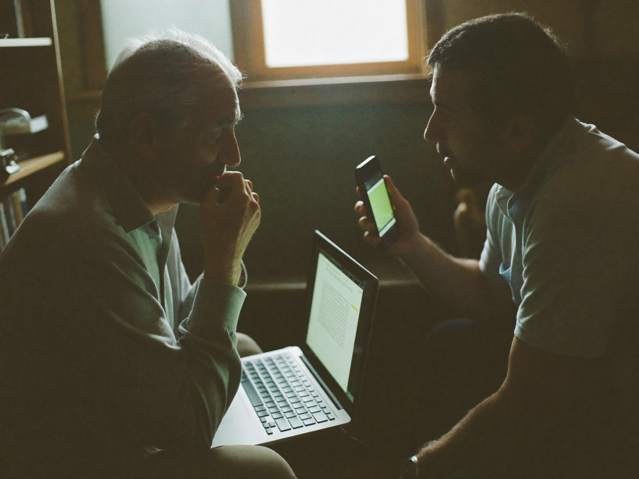 Two pastors talking face to face, one with a laptop open on his knees, the other holding up a phone