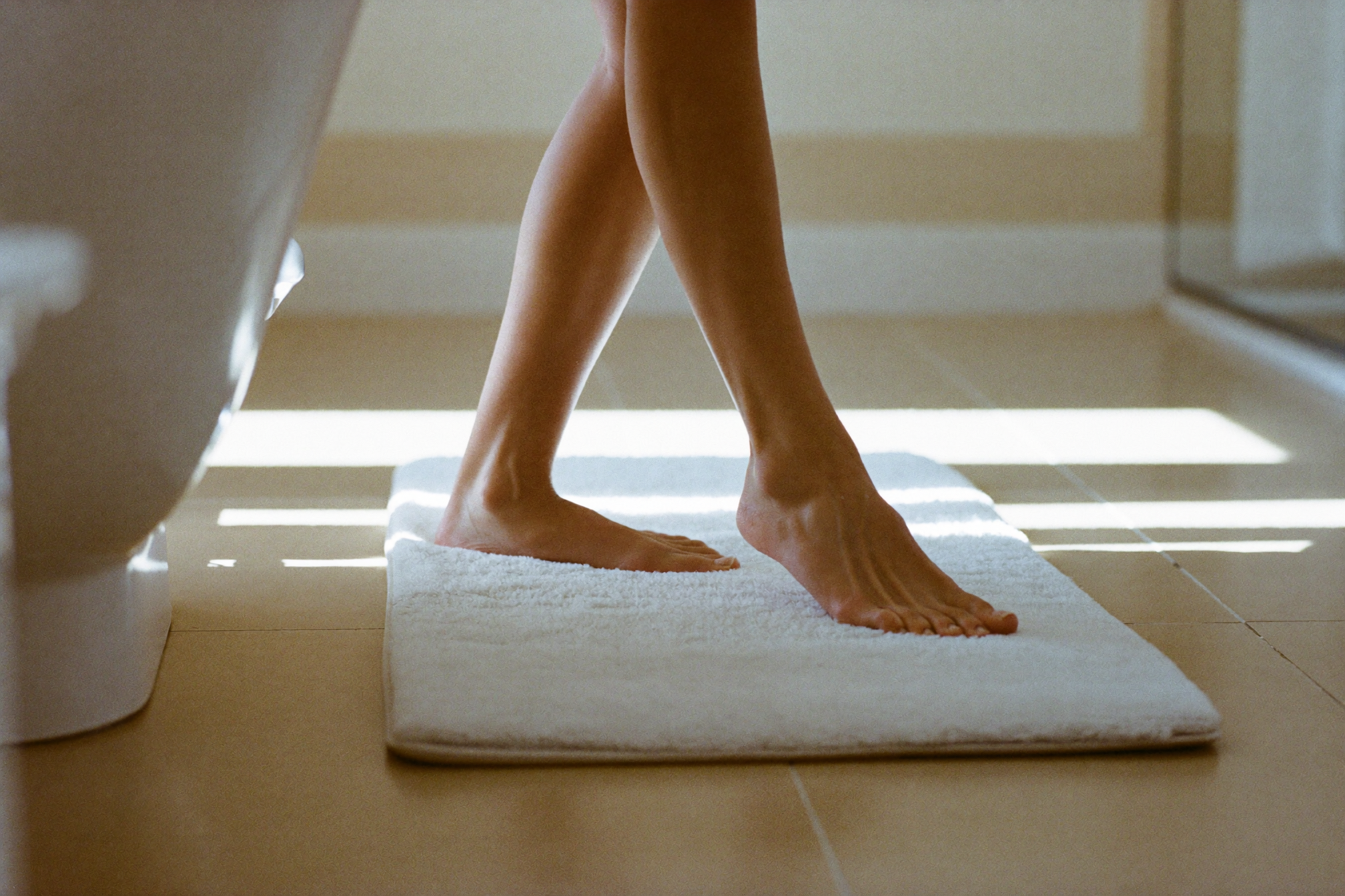 Bare feet resting on a soft bathmat in a sunlit, minimal bathroom