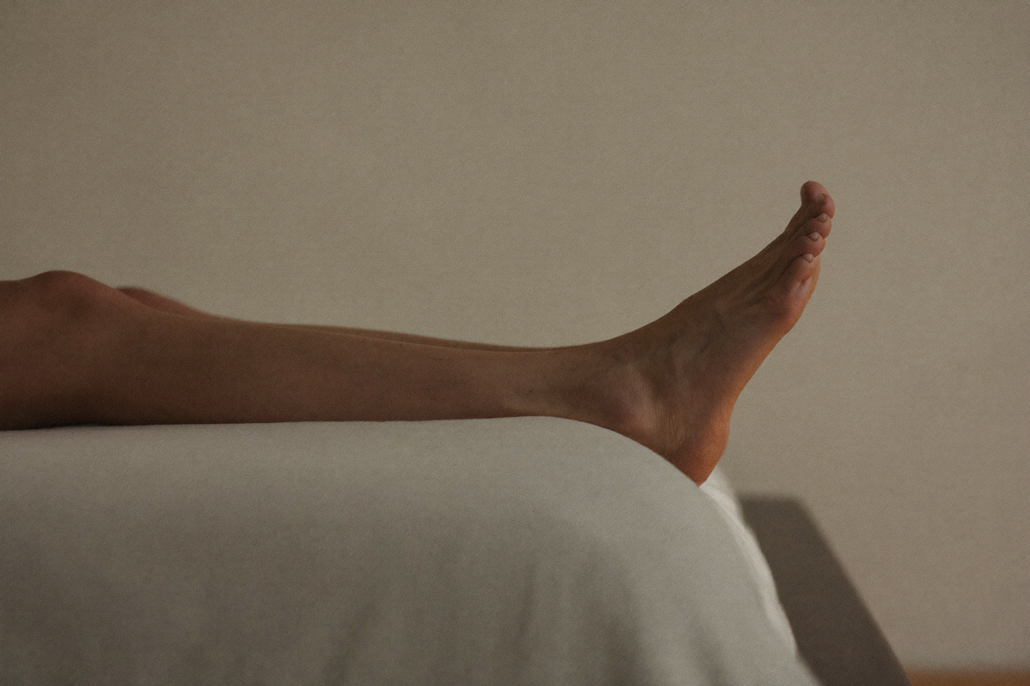 Person gently stretching one bare foot while seated on the edge of a bed in soft evening light