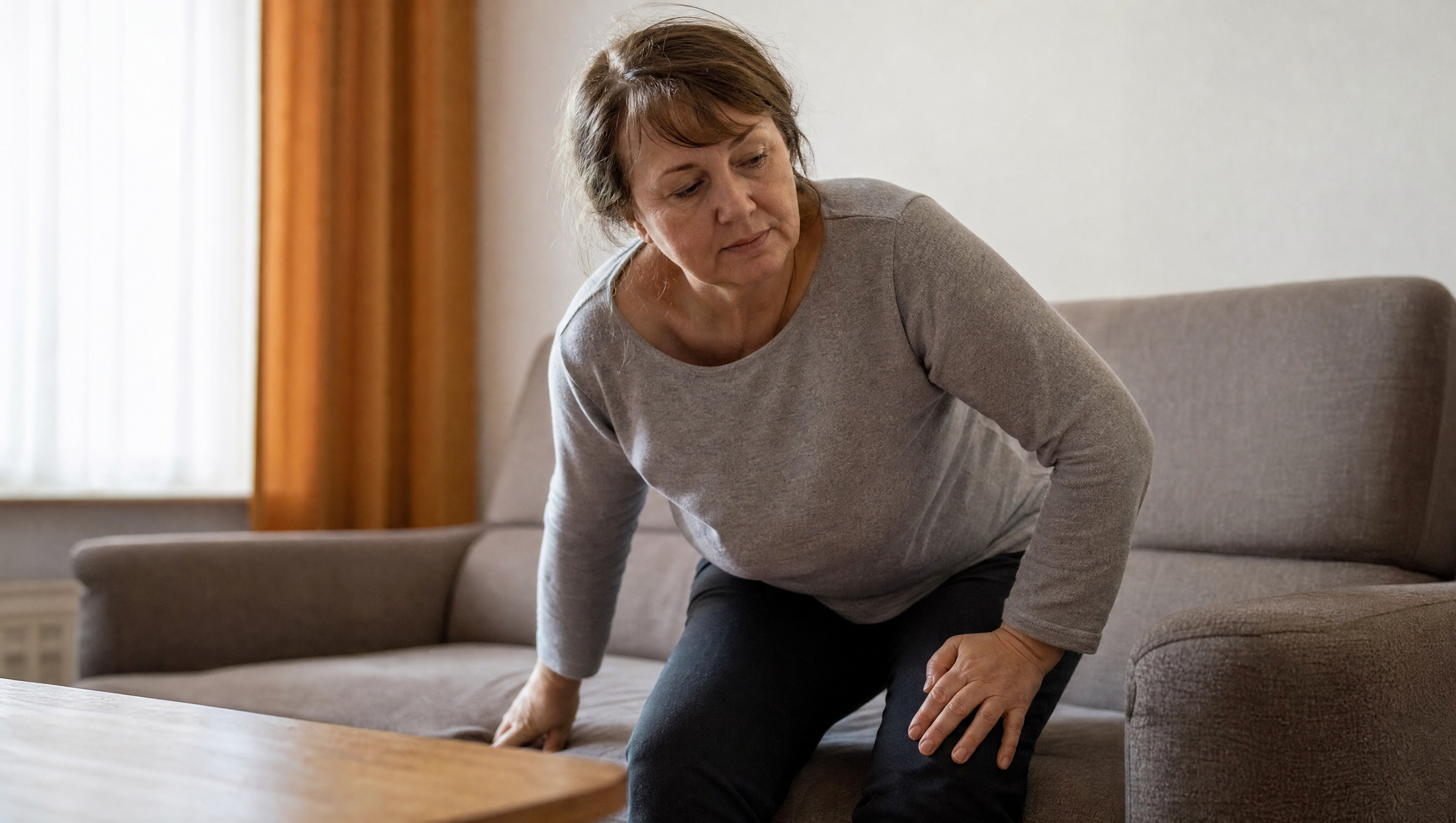 Frau um die 55 stützt sich beim Aufstehen vom Sofa ab, Morgenlicht