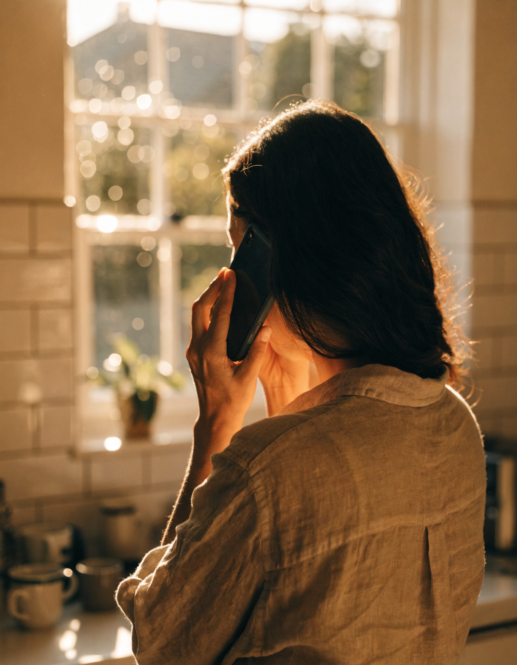 A woman standing in her kitchen, seen from behind, pressing her phone to her ear with one hand and covering her mouth with the other — overwhelmed with relief at the news from her clinic.