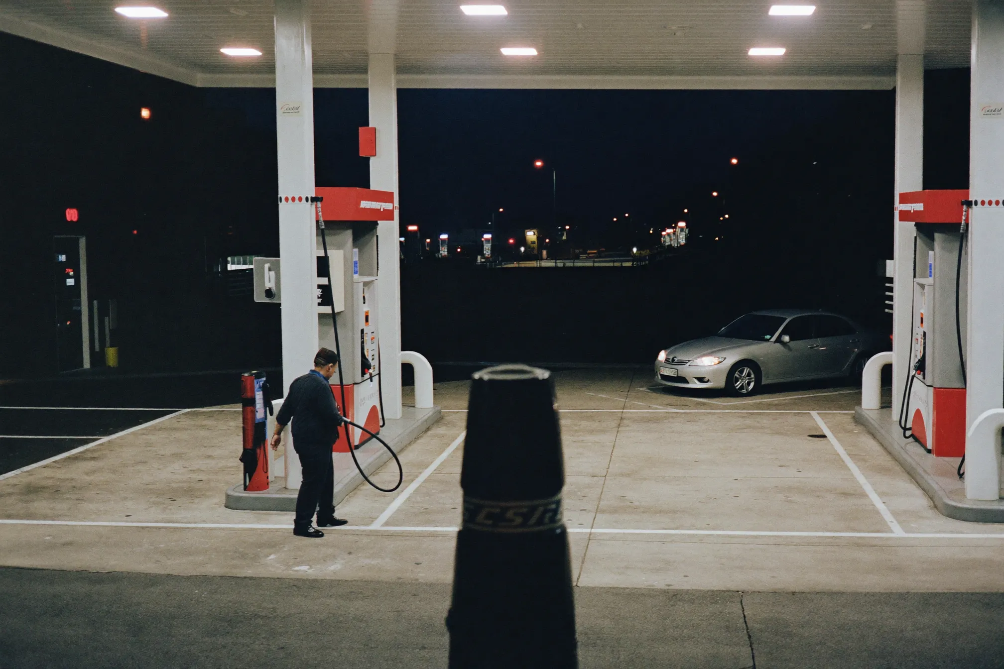 A gas station forecourt at night with an attendant between the pumps
