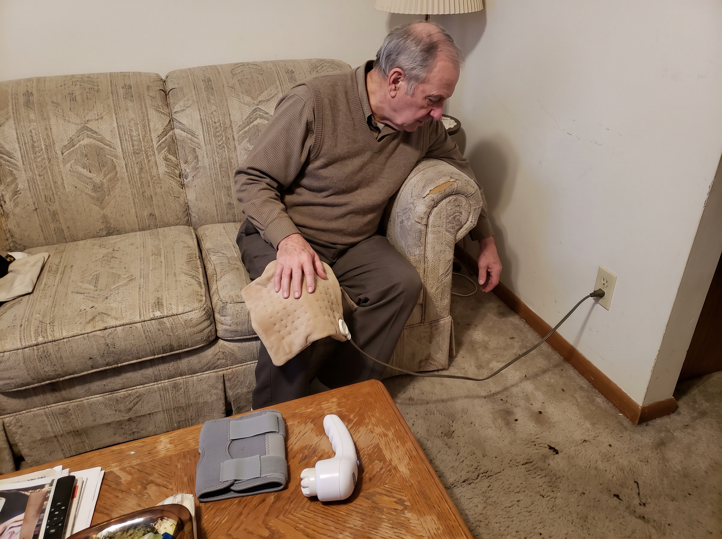 A man holding a corded heating pad on his knee, its cord stretched across the carpet to the wall outlet