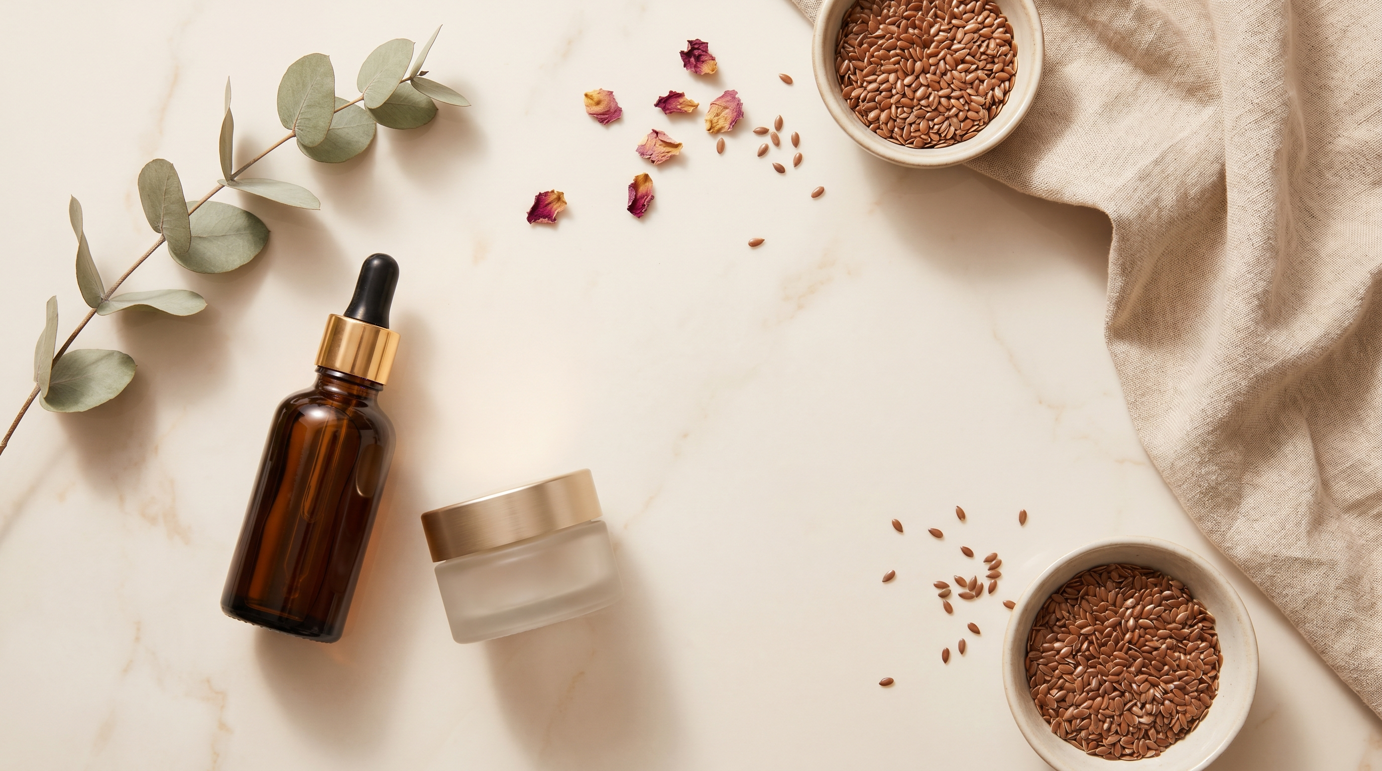 Composition de soins naturels anti-âge : sérum ambré, crème, pétales séchés et feuilles vertes sur marbre crème