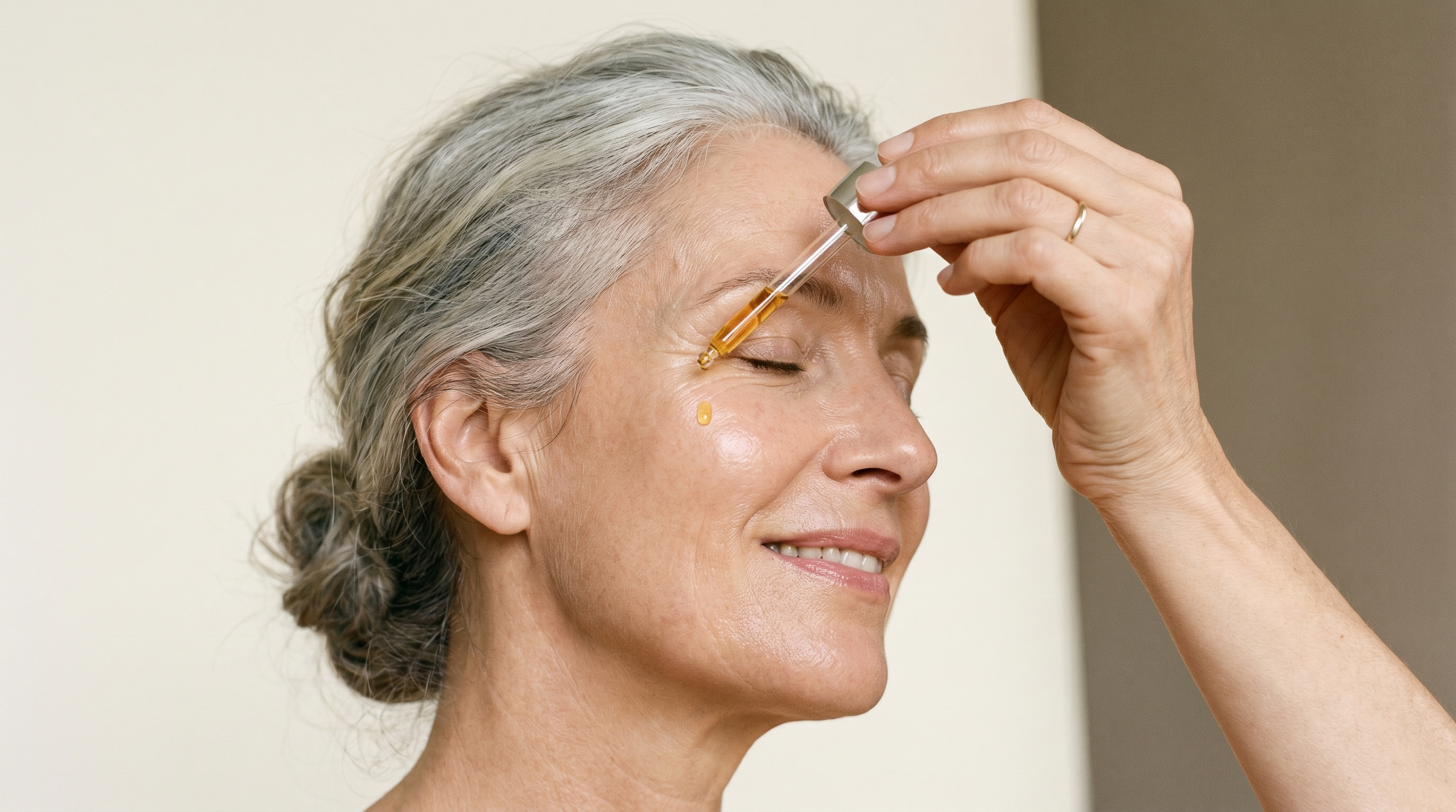 Femme élégante de 55 ans tient une pipette en verre et applique un sérum vitamine C sur sa joue, lumière douce