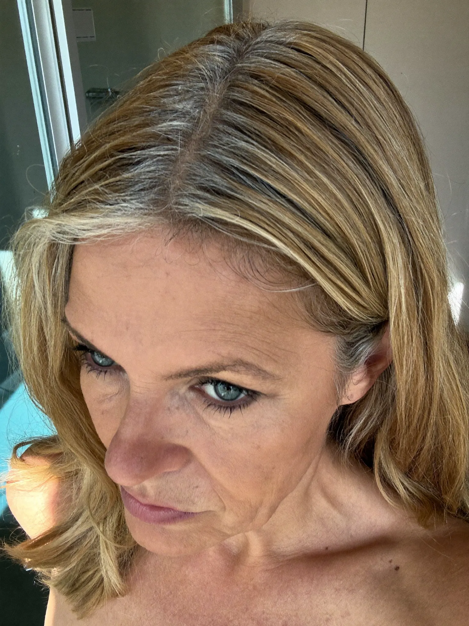 Woman noticing first gray regrowth around her hair part in a bathroom mirror