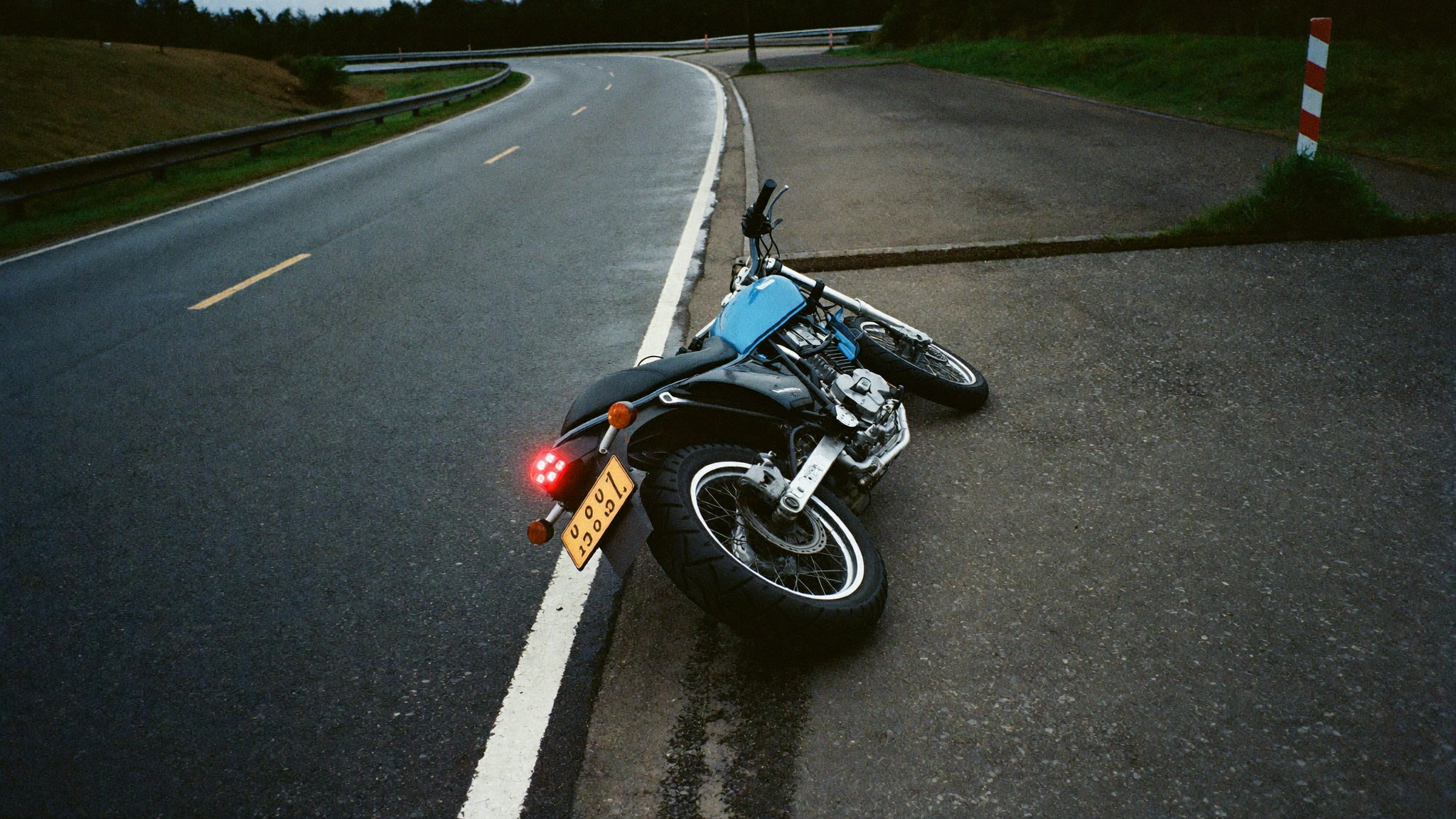 A commuter motorcycle down on its side on a wet road at dusk, tail light glowing