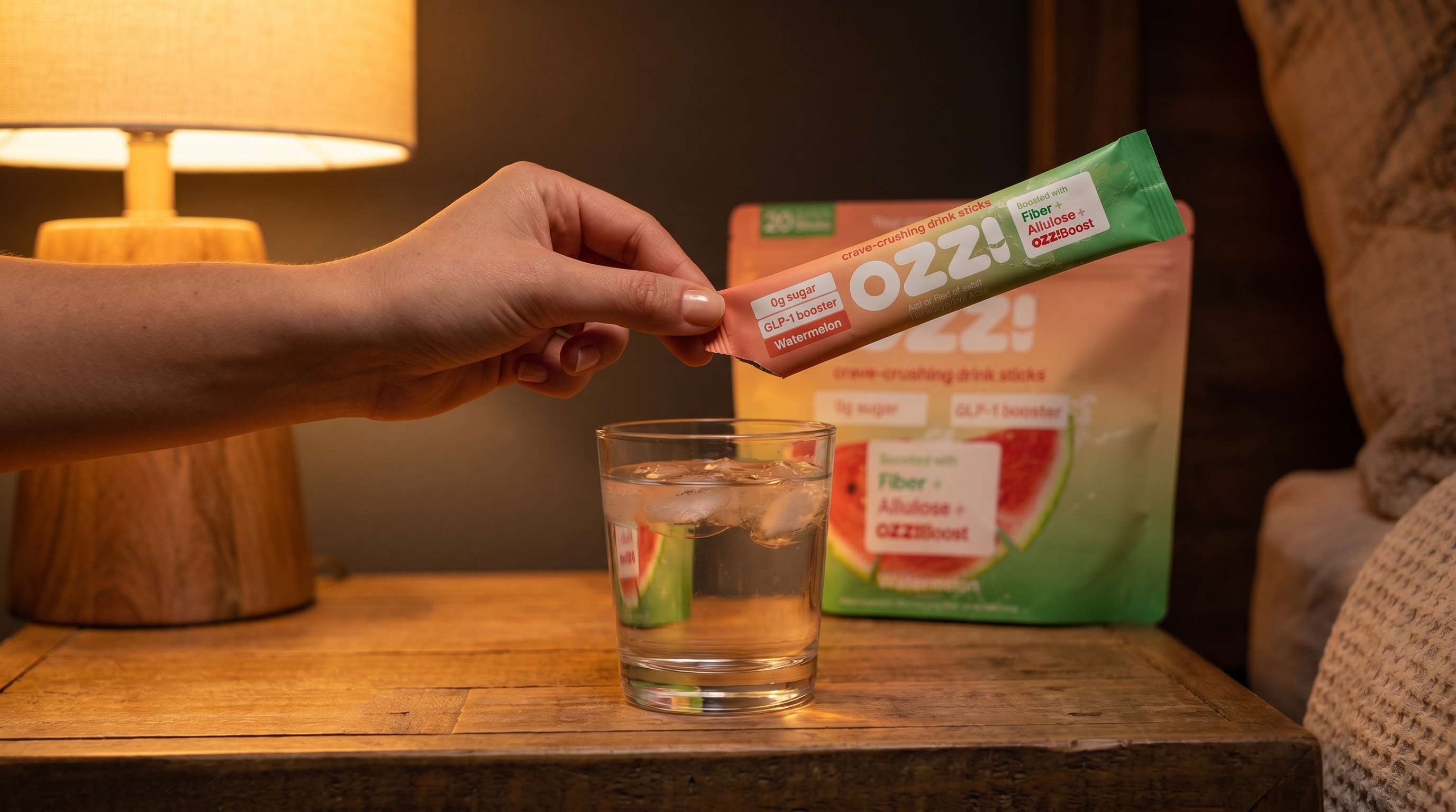 Hand tearing open an Ozzi watermelon drink stick over a glass of water on a nightstand at night