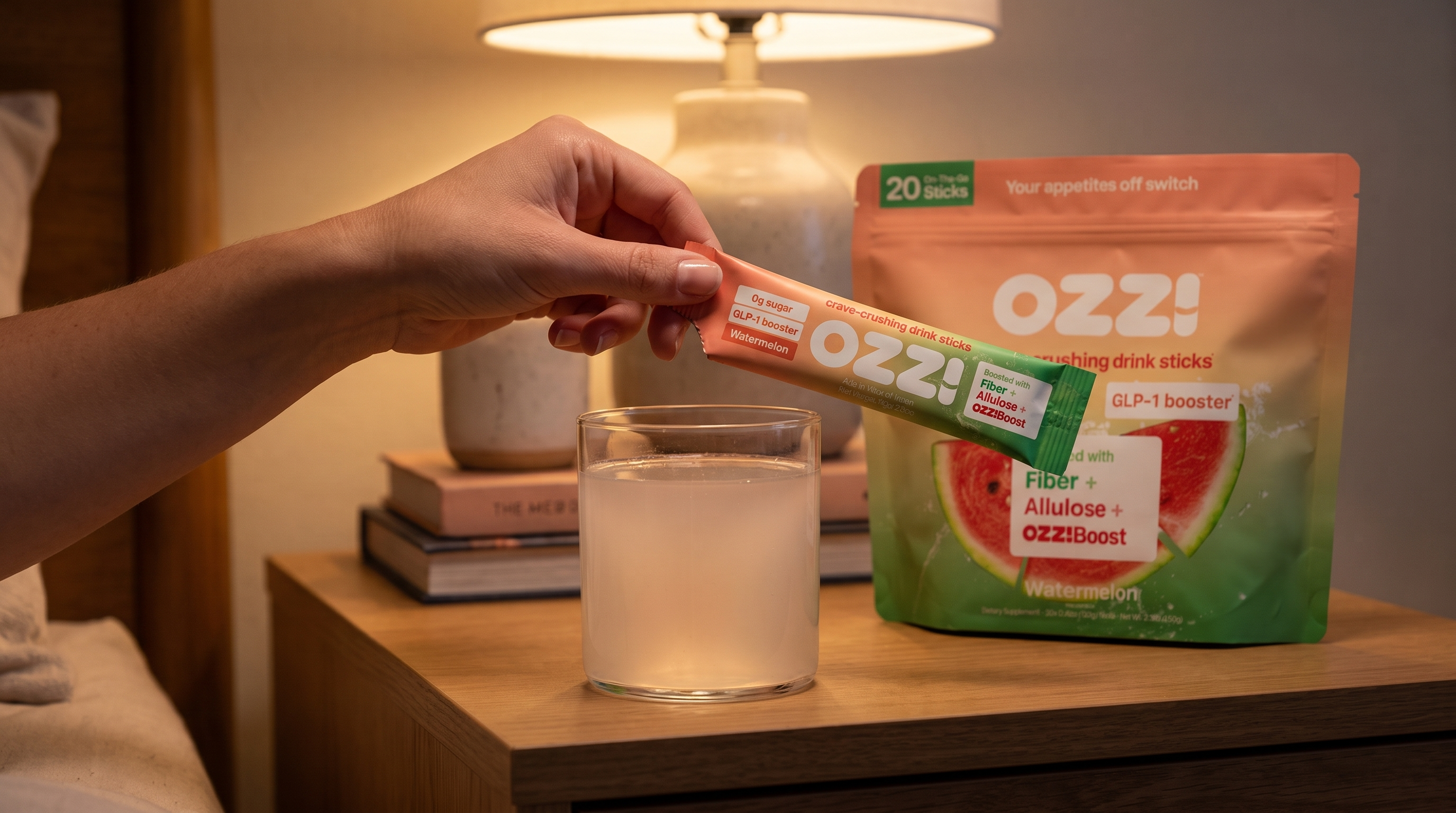 A hand tearing open an Ozzi watermelon stick over a glass of water on a nightstand in evening light
