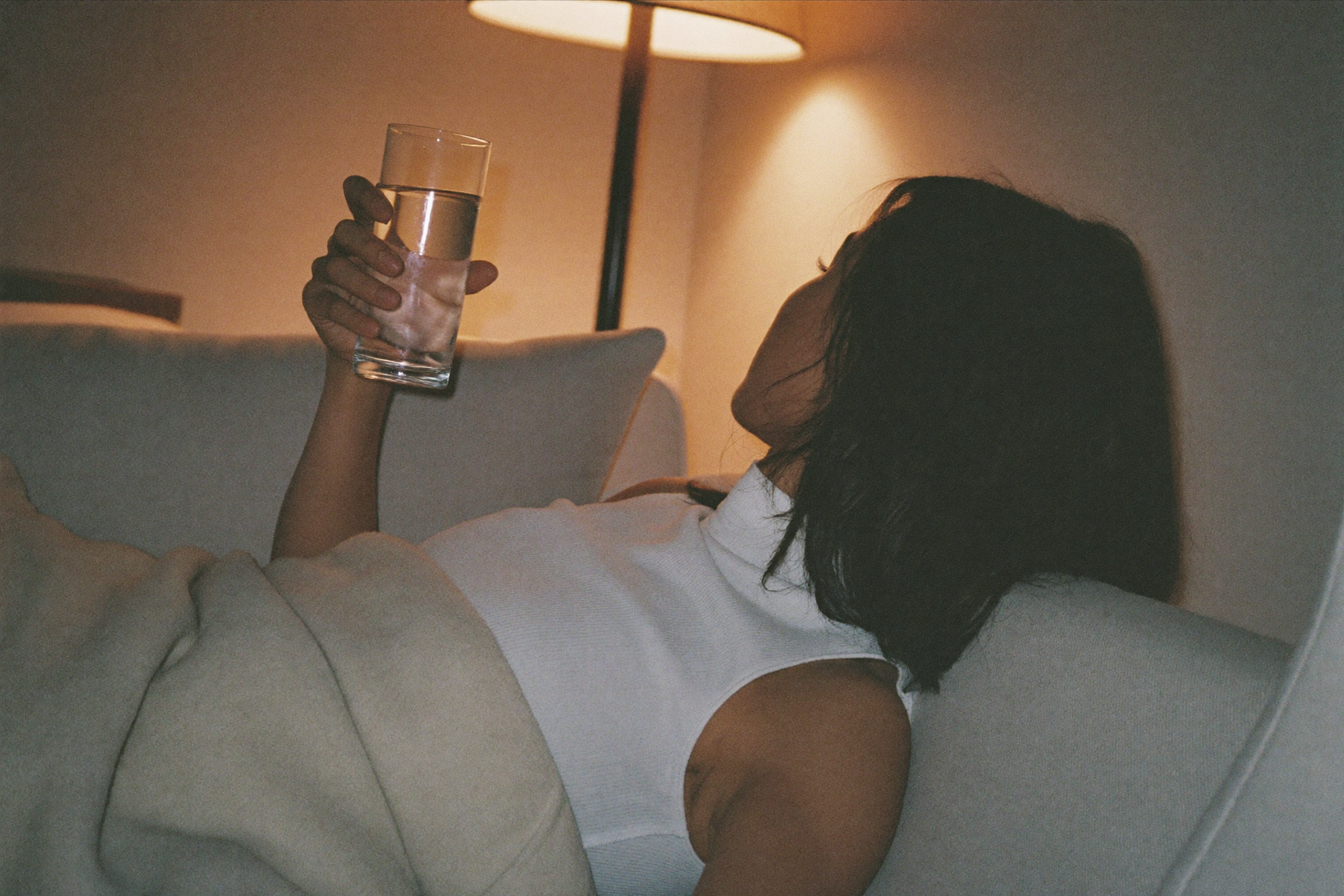 Woman relaxing on a couch in the evening holding a glass of water, warm lamp light
