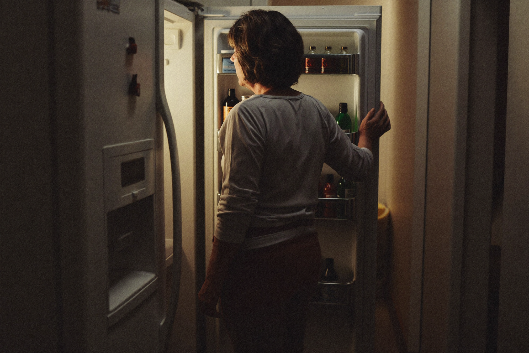 A woman standing at an open refrigerator in a dark kitchen at night