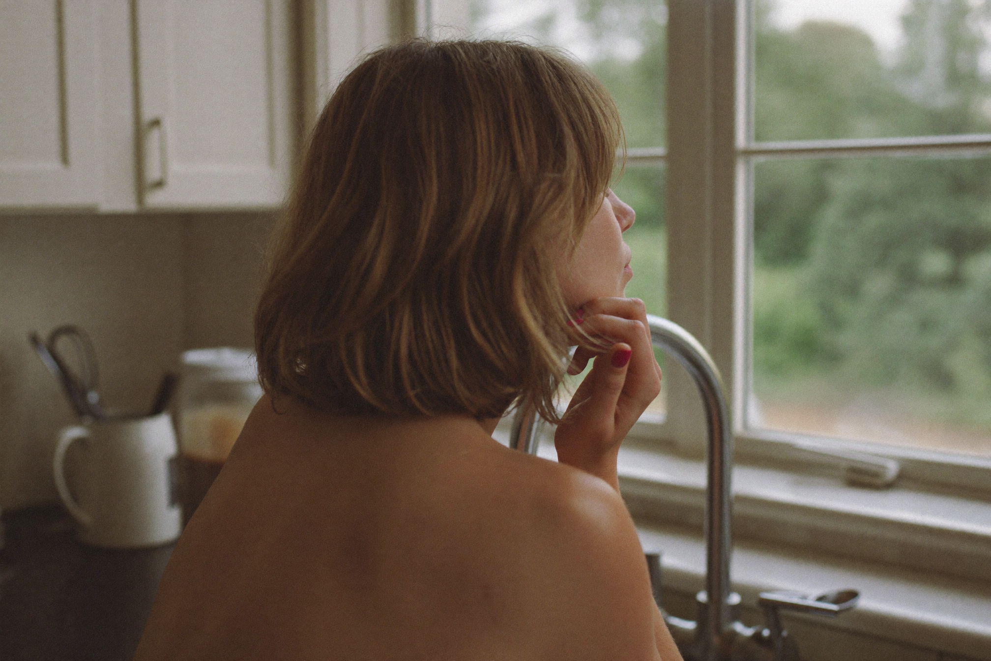 A woman standing in a calm sunlit kitchen, looking out the window