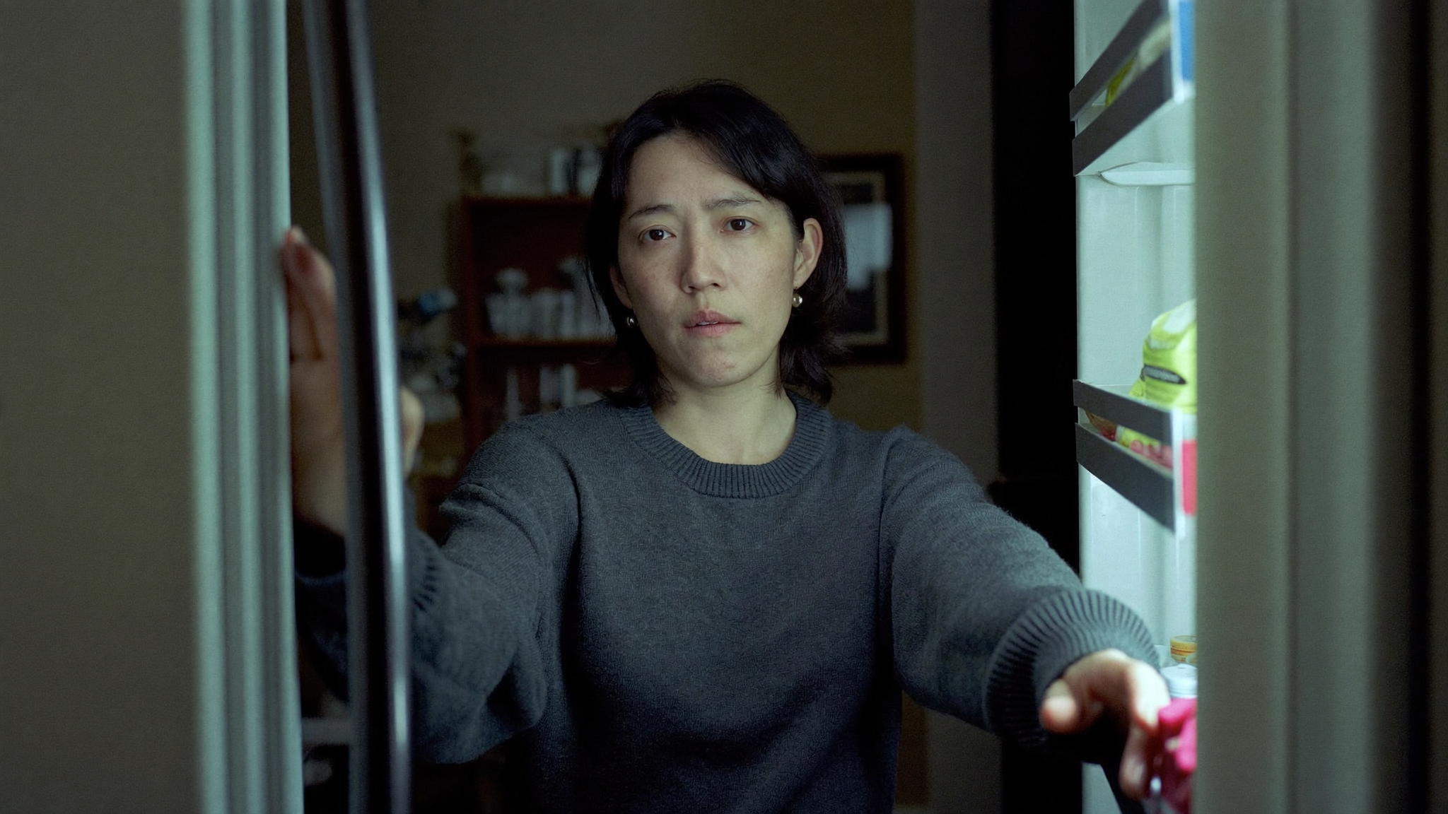 A woman in her forties standing at an open refrigerator in a dim kitchen late at night, looking tired and conflicted.