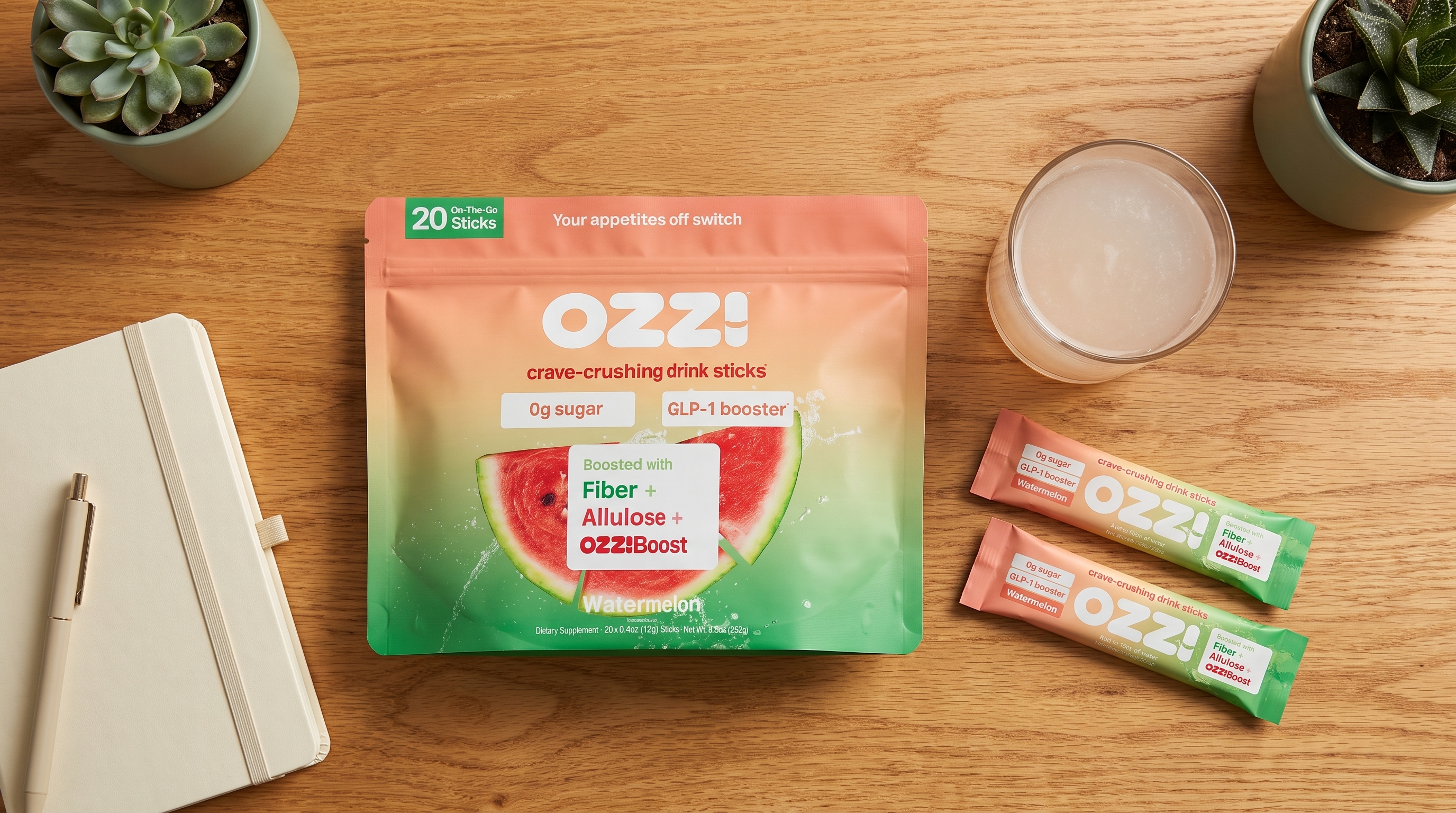 Overhead flatlay of the Ozzi watermelon pouch, two stick packs, and a glass of the prepared drink on a wooden desk beside a notebook.