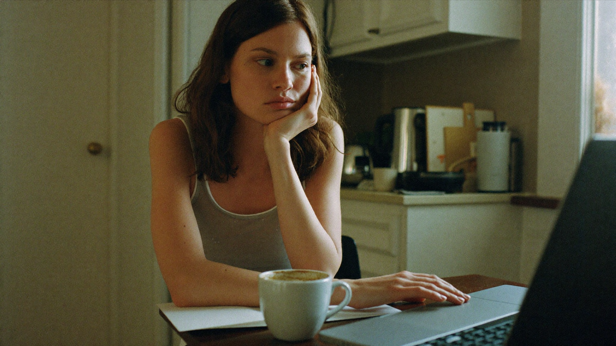 A woman in her late thirties sitting at a kitchen table in mid-afternoon light, chin on hand, staring past a half-finished mug of coffee