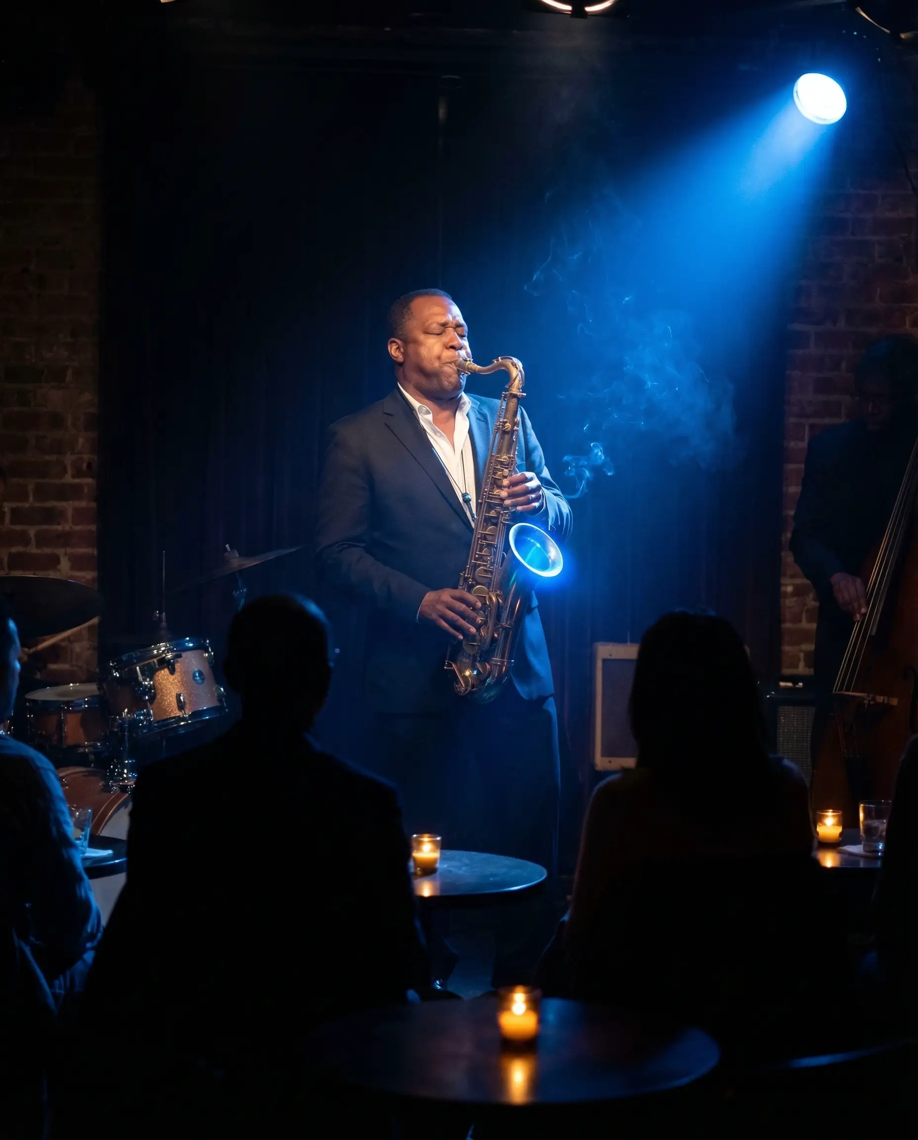 SHOT: Medium shot from behind the audience, looking toward a small stage. A saxophone player stands in a single blue spot. Cigarette smoke drifts through the light beam.

SUBJECT
A saxophone player — a man in his fifties, African American, wearing a dark suit, white shirt with open collar, no tie. He plays tenor saxophone — the brass catches the blue stage light as a bright curved highlight along the bell. His eyes are closed. His cheeks puffed slightly with the embouchure. His fingers work the keys — dark shapes against bright brass.
Expression: Eyes closed, transported — not performing for the audience, inside the music. Body leans slightly back, weight on heels.

SCENE
A small jazz club — capacity 60. Small round tables with candles in the dark foreground. The audience visible as dark shapes and occasional candle-lit faces. The stage is tiny — barely room for a quartet. Drum kit and upright bass visible at stage edges, slightly out of the spot's reach. Exposed brick walls. Low ceiling with acoustic tiles.

CINEMATOGRAPHY
Lighting: One blue-gelled spot from above-right — tight, hitting only the saxophonist. The blue light catches the brass, his face, and the rising smoke. Candles on tables provide tiny warm amber points in the dark audience area. Contrast: cold blue stage, warm amber audience. Smoke from the crowd drifts through the blue spot beam — visible as slow-moving grey-blue wisps.
Tone: Blue spot on stage, warm amber candle points in darkness.

STYLE
Blue Note Records album cover aesthetic — the musician in blue light.
No text, no signage, no writing, no logos, no watermarks.