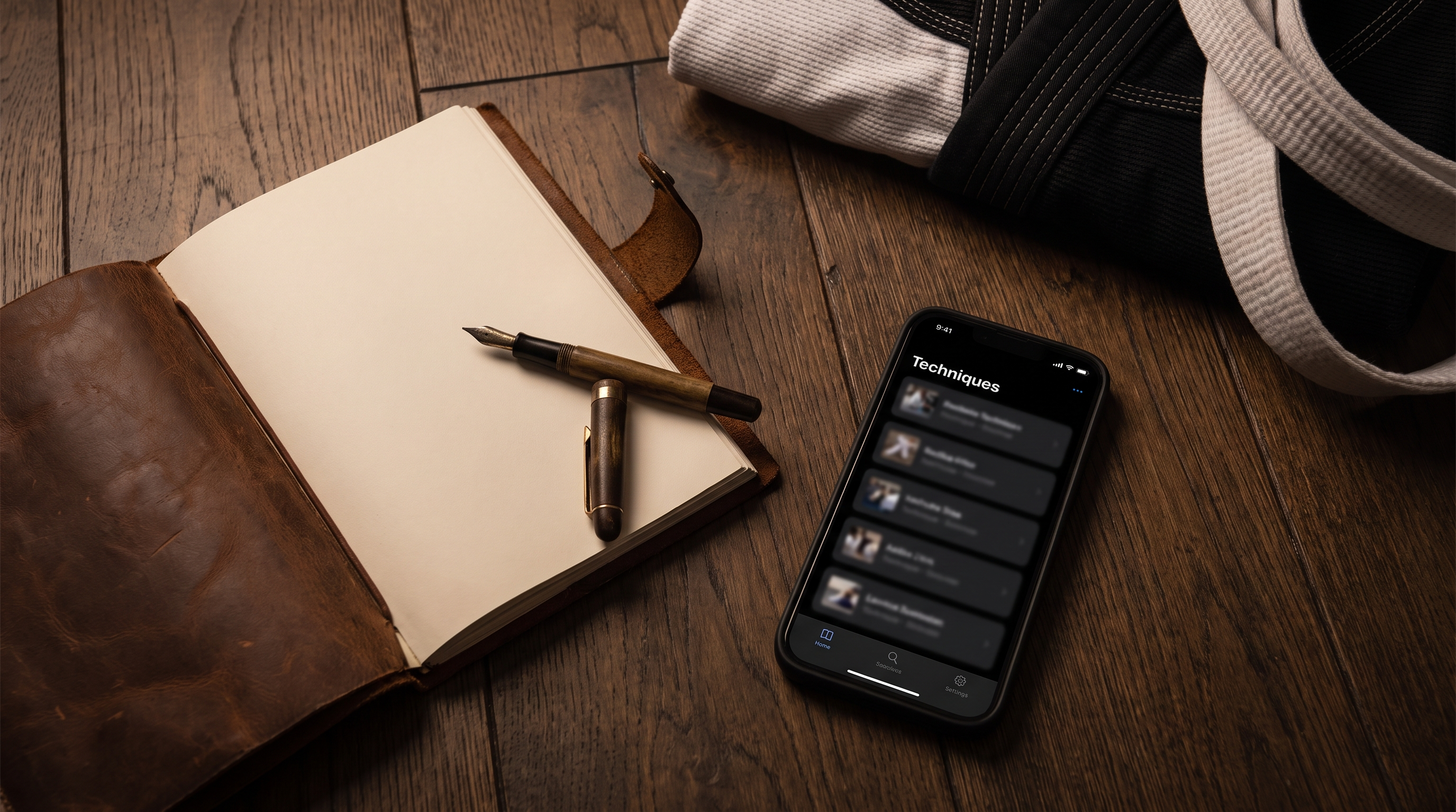 Overhead view of an open BJJ notebook with a pen next to a smartphone showing a technique card, on dark hardwood with a folded gi.