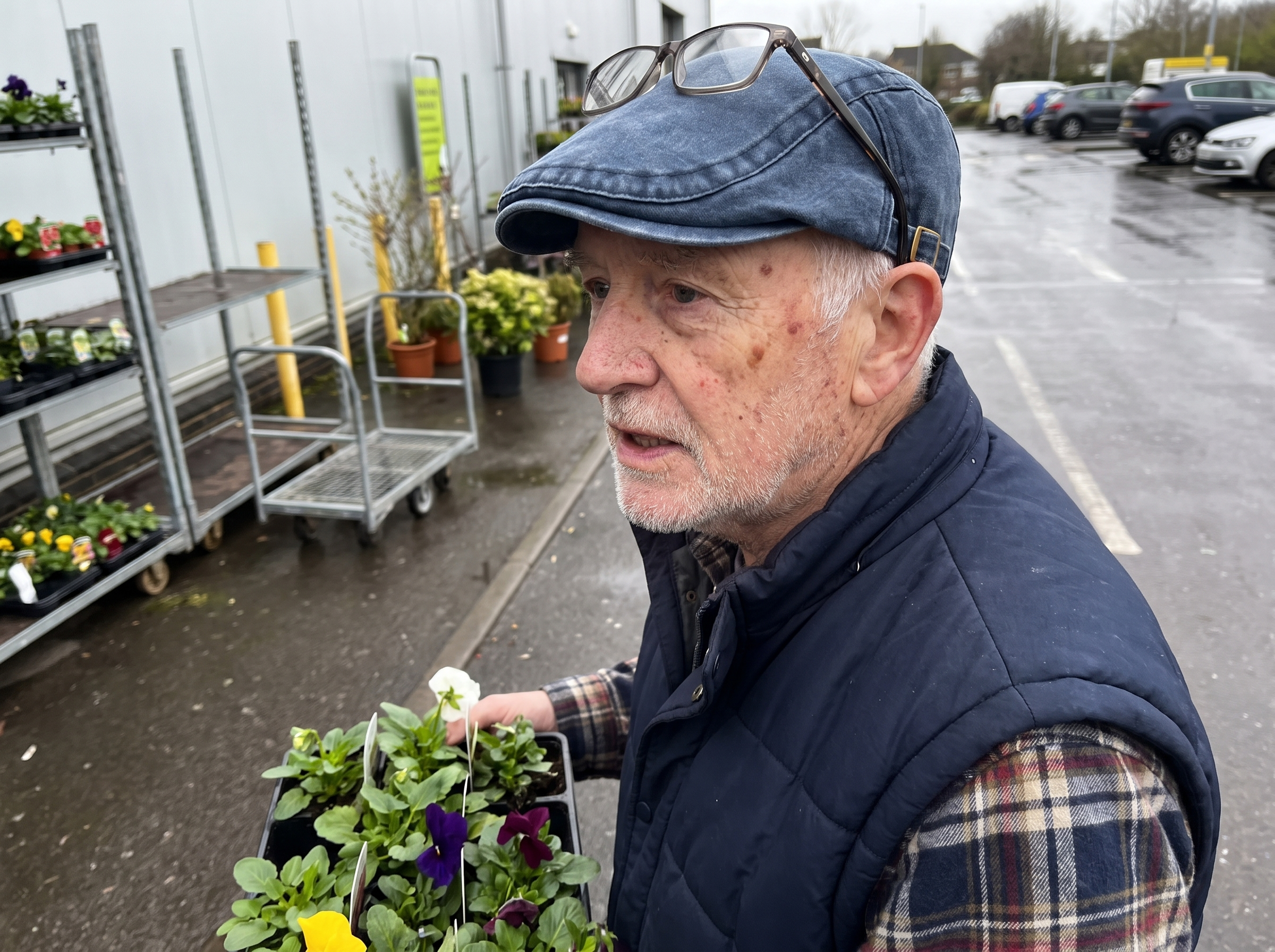 A man in his seventies at a garden centre holding a tray of plants, wearing the denim flat cap