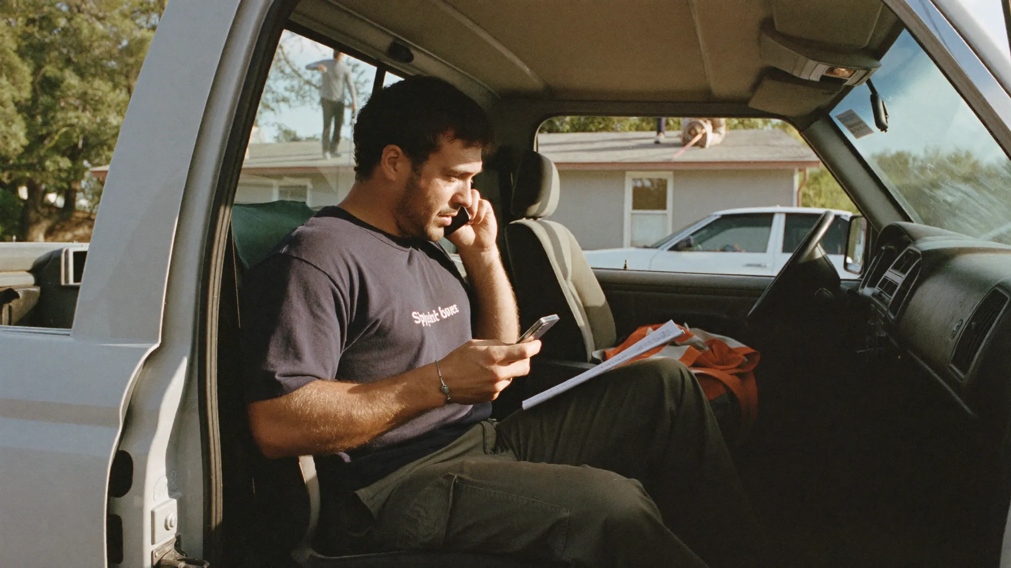 A roofing contractor sitting in the open door of his work truck updating a job on his phone with a paper estimate on his lap