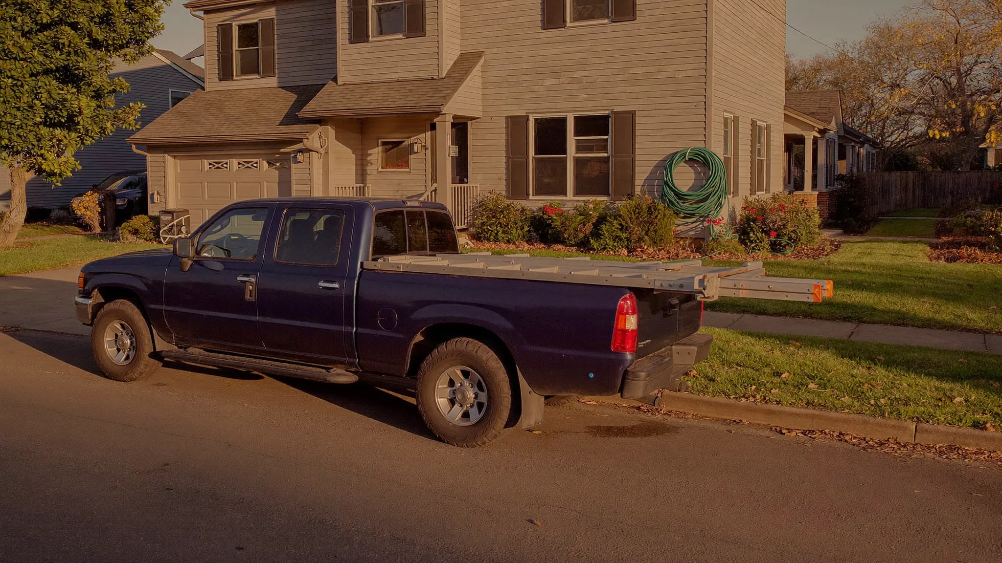 A navy work pickup with an extension ladder in the bed parked at the curb of a quiet suburban street at golden hour