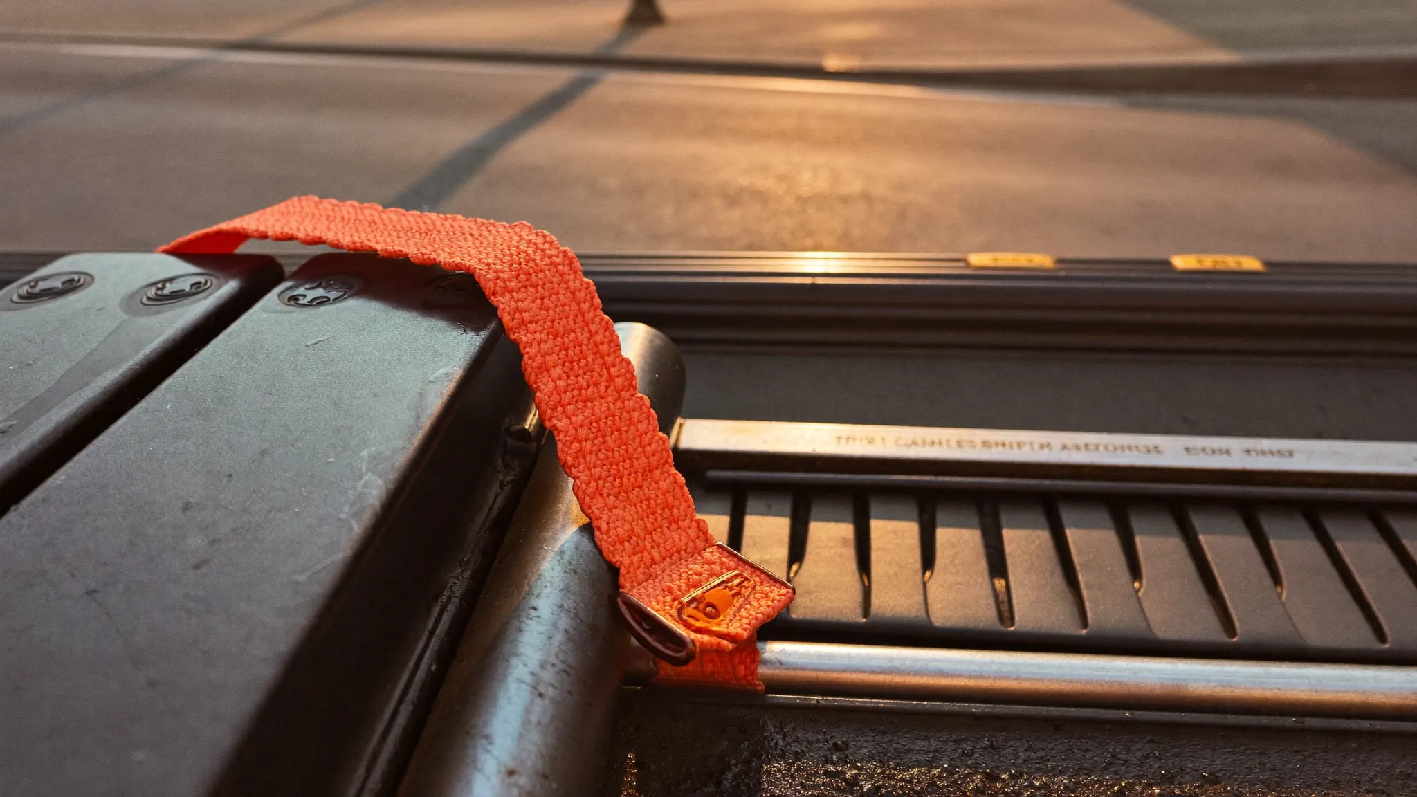 A bright orange ratchet strap cinched over the steel track of a compact machine on a black trailer deck in morning light