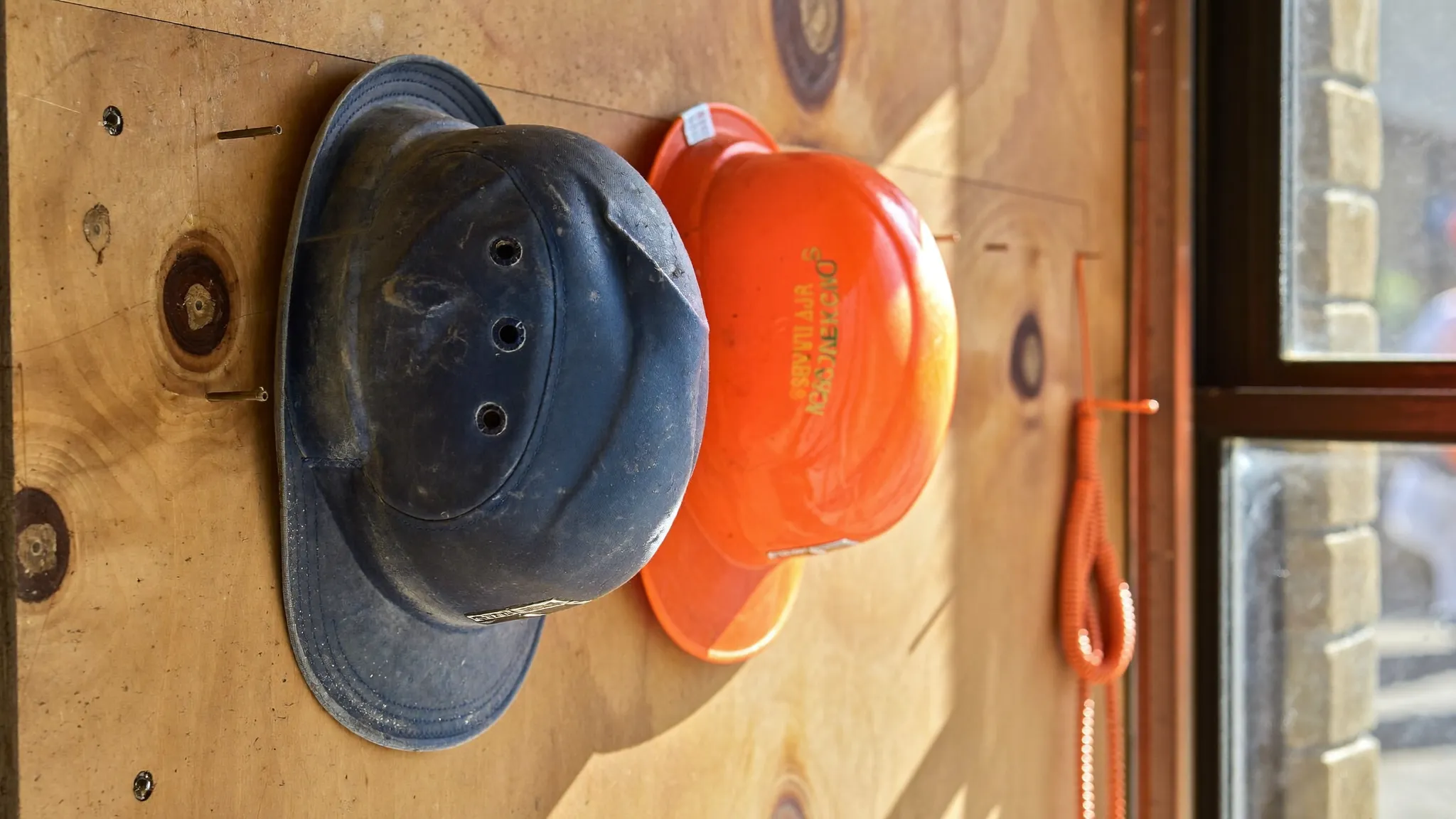 A worn navy hard hat and a brand-new orange hard hat hanging side by side on a plywood shop wall