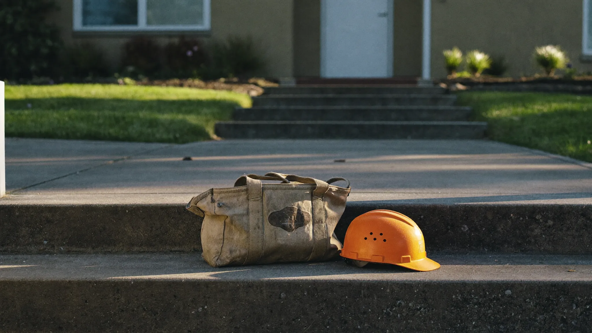 A worn canvas tool bag and an orange hard hat resting on the concrete front steps of a house in early morning