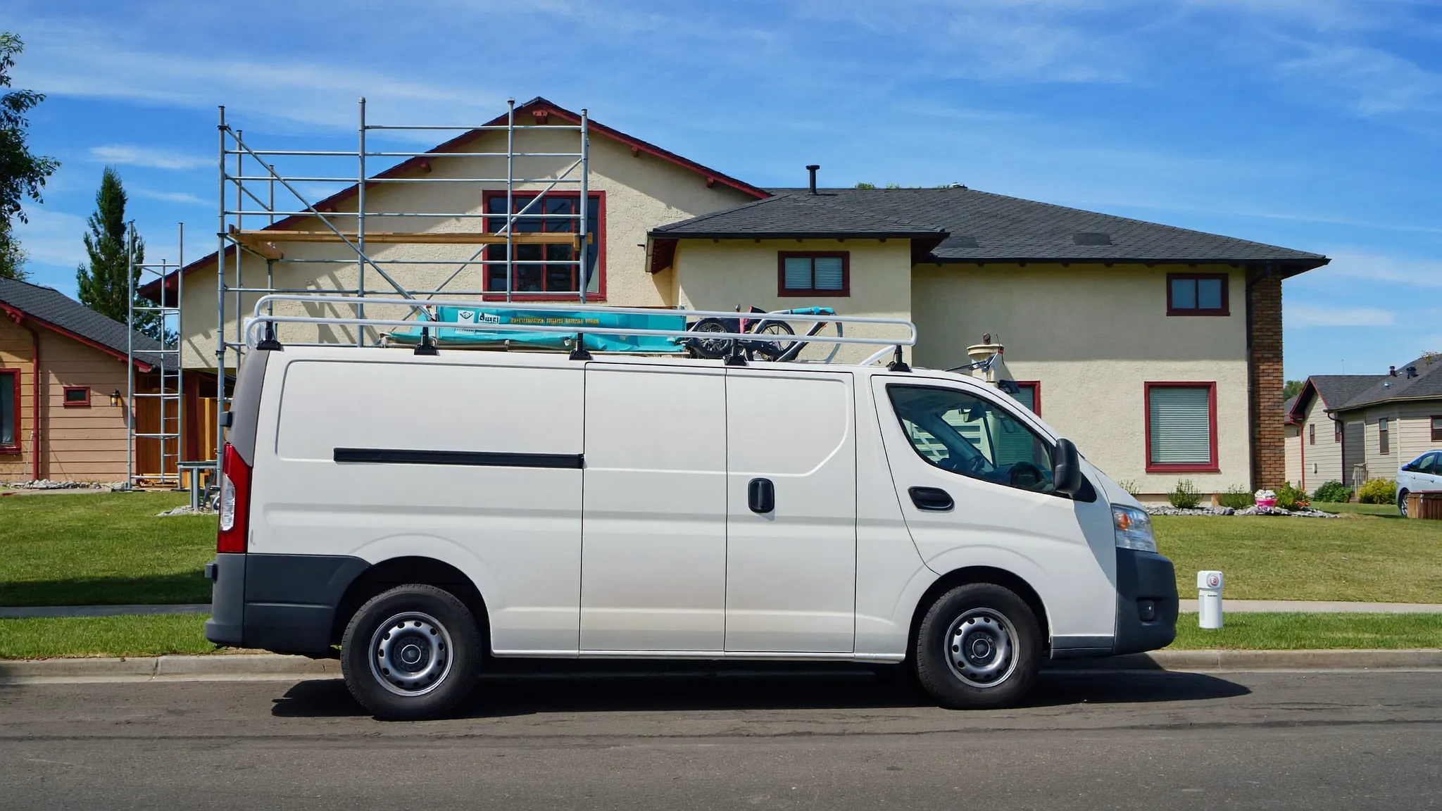 A plain white work van with a ladder rack parked outside a suburban house under renovation with scaffolding