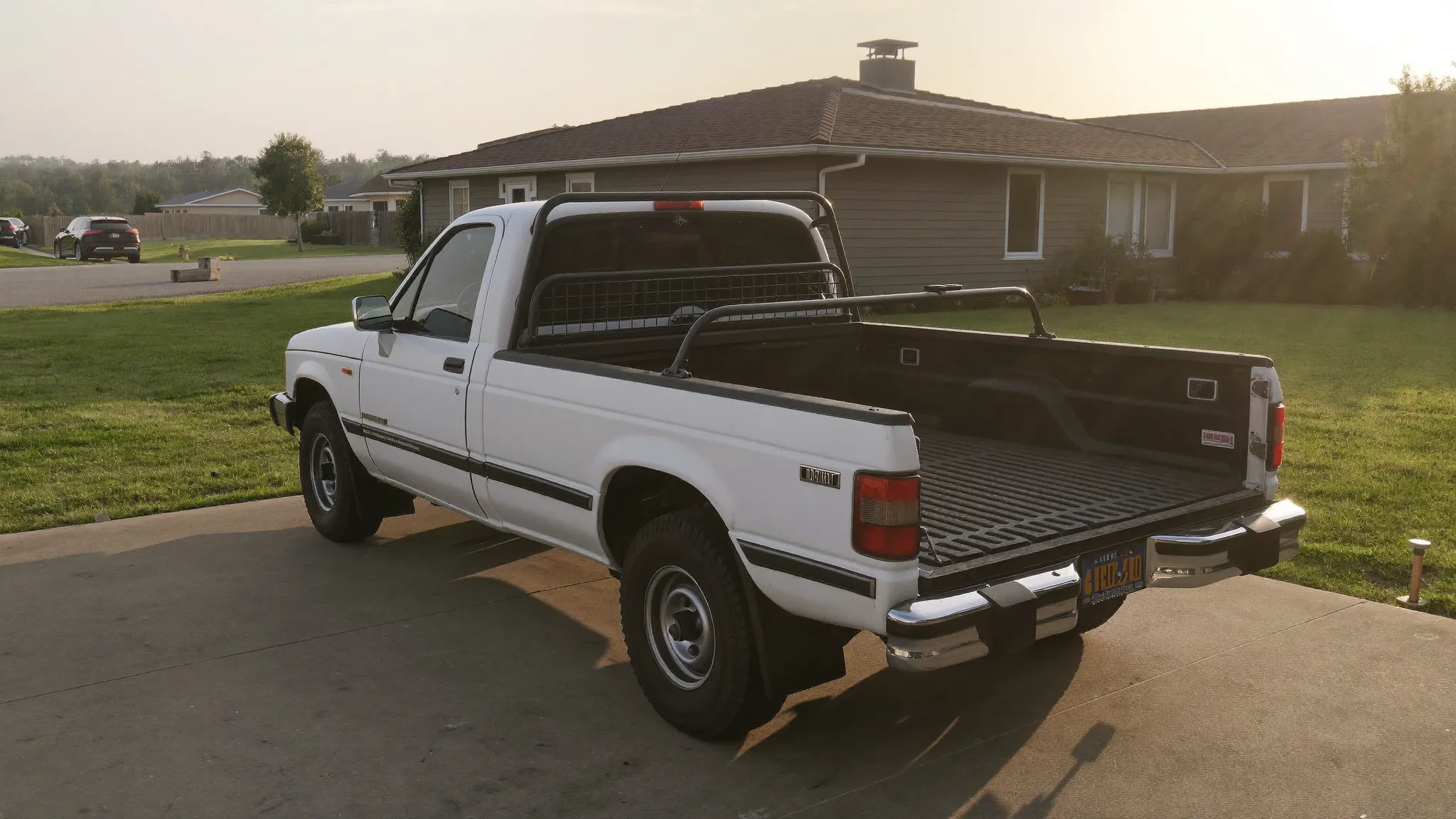 A plain white work pickup truck parked on a suburban driveway at golden hour with the tailgate down