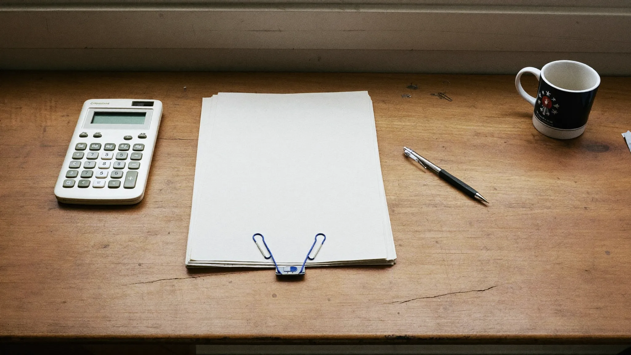 A stack of paperwork on a clipboard beside a calculator, a pen, and a coffee mug on a worn wooden desk