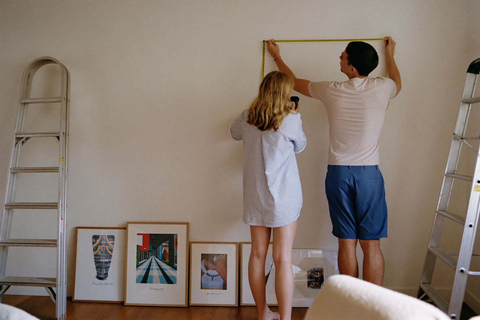 Couple measuring their living room wall with framed prints leaning against the wall