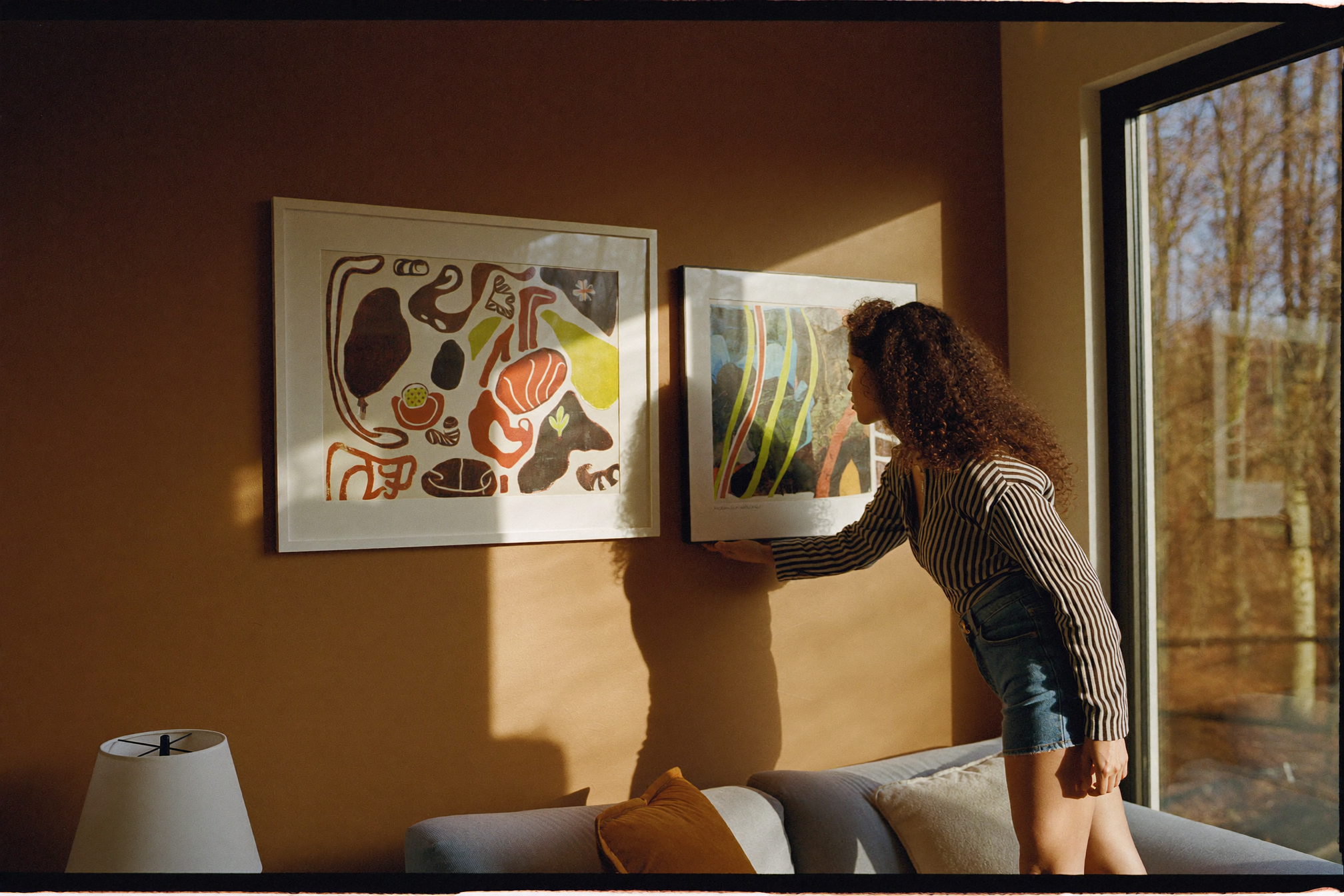 Woman hanging a framed art print on a warm terracotta living room wall in autumn light