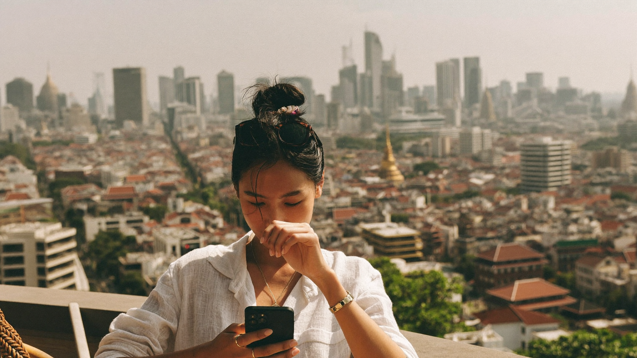 Traveller checking a smartphone at a sunlit rooftop terrace overlooking Bangkok's city skyline