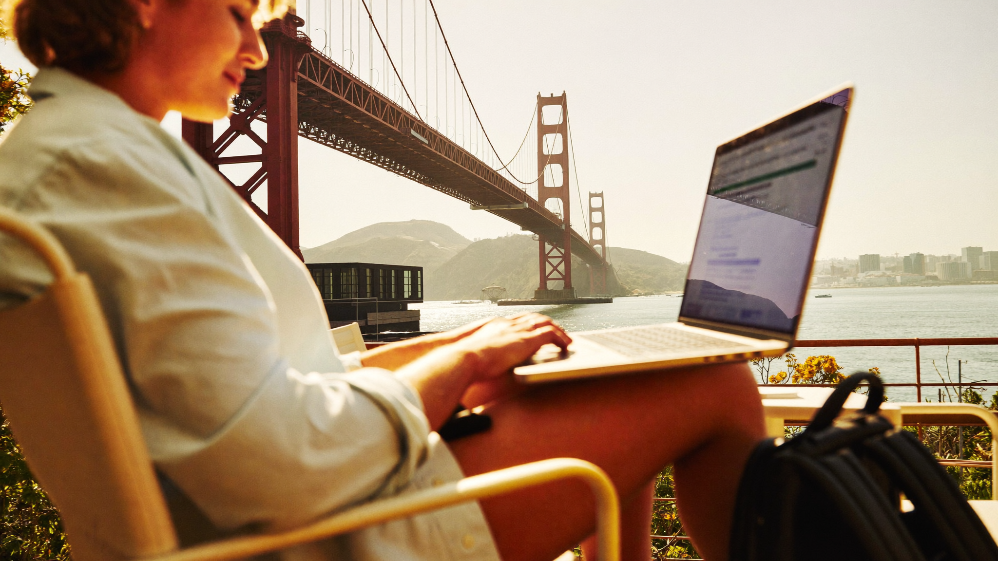 Solo traveller sitting at a San Francisco rooftop café, laptop open, bay and bridge visible in the background