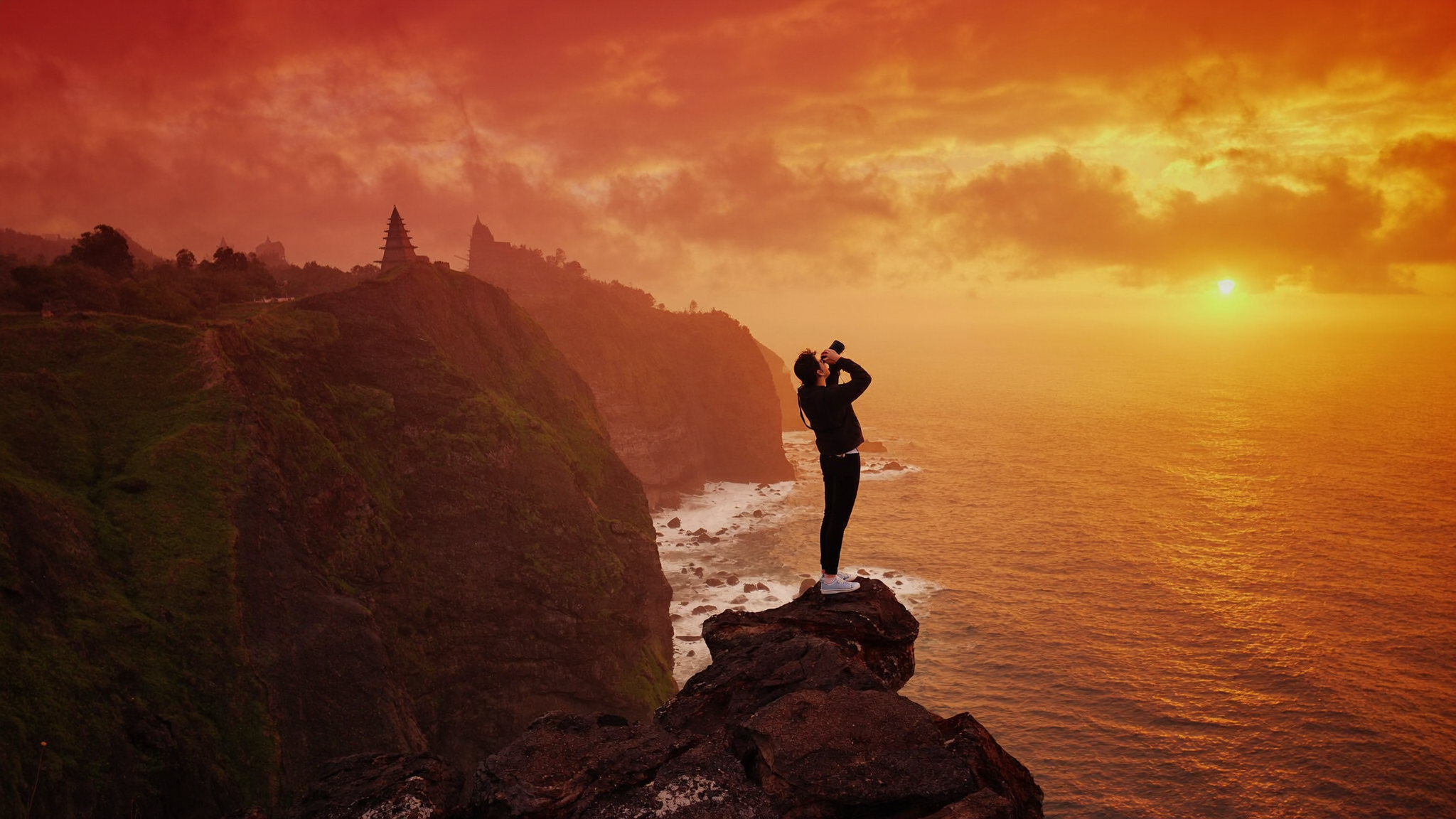 Traveller photographing a dramatic Bali clifftop sunset at Uluwatu, ocean crashing against rocks far below