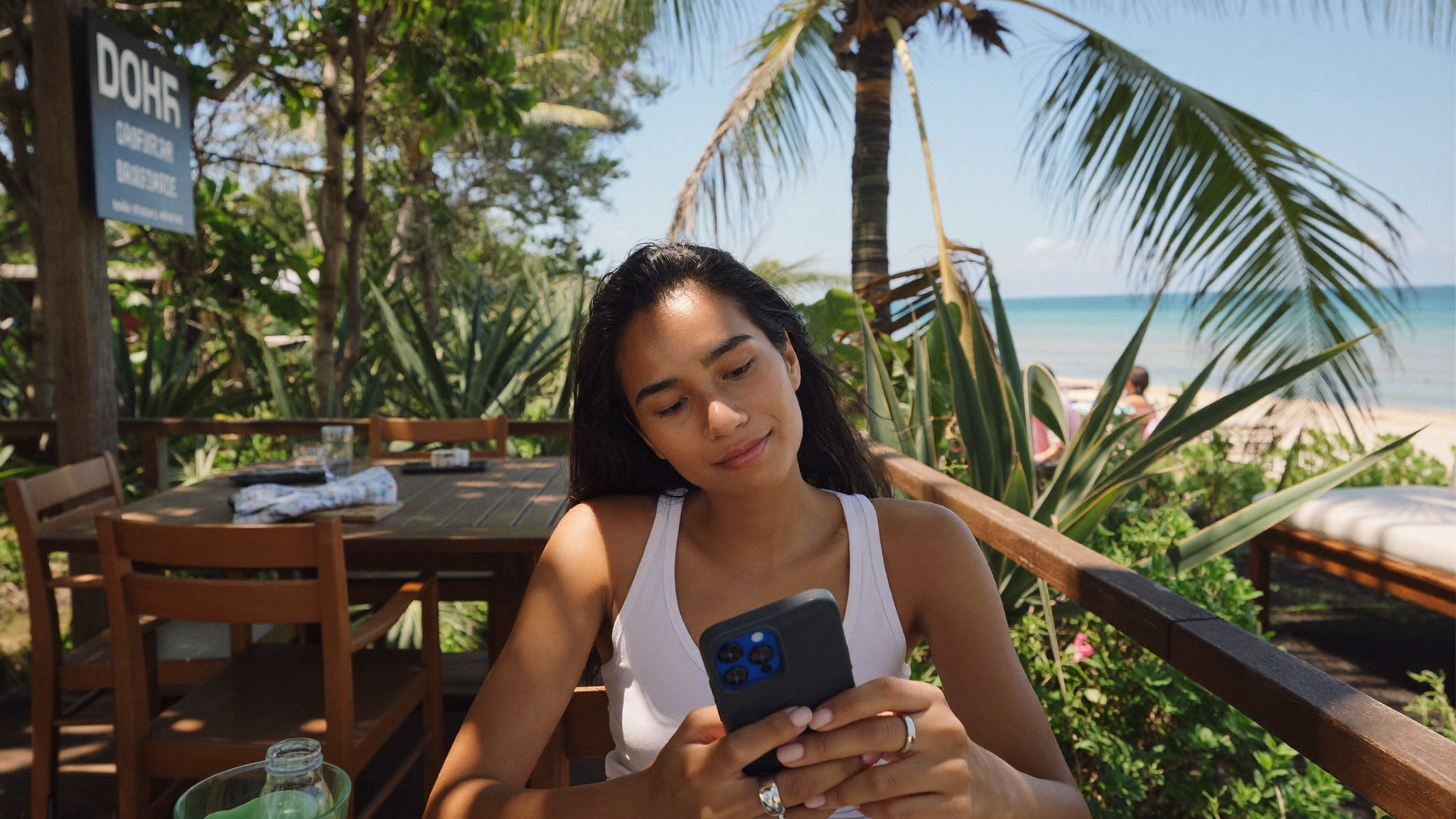 Traveller sitting at a Bali beach café terrace, smartphone in hand, turquoise ocean stretching to the horizon