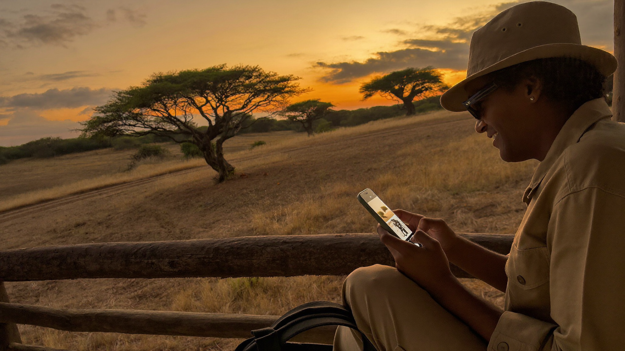 Traveller in safari clothing using a smartphone at a lodge terrace, acacia trees and savannah visible at dusk