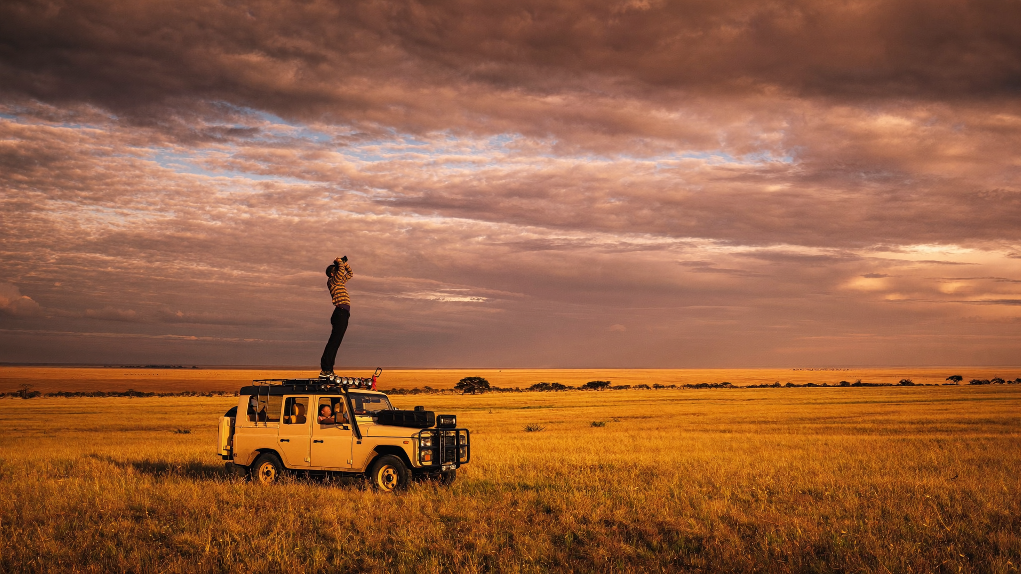 Solo traveller with binoculars standing at the edge of a safari vehicle, vast Kenyan savannah stretching to the horizon