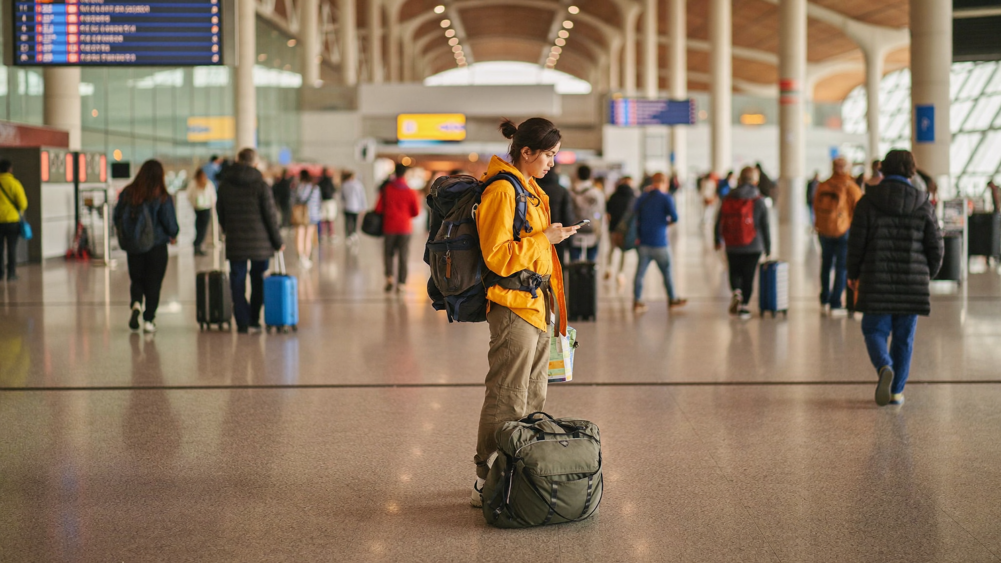 Backpacker at an airport departure hall, luggage at feet, checking phone before boarding