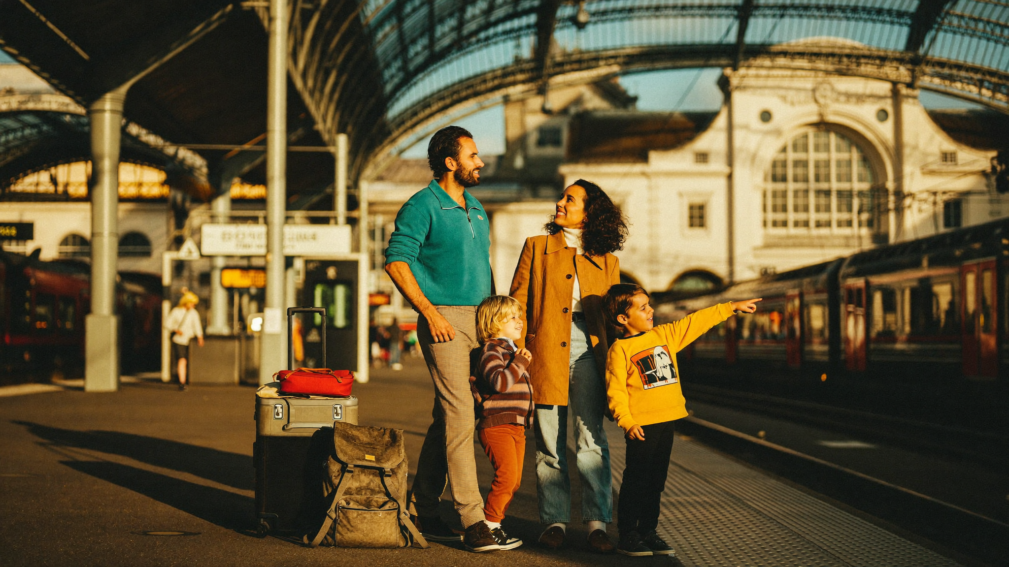 Family at a sunlit European train station platform, luggage stacked beside them