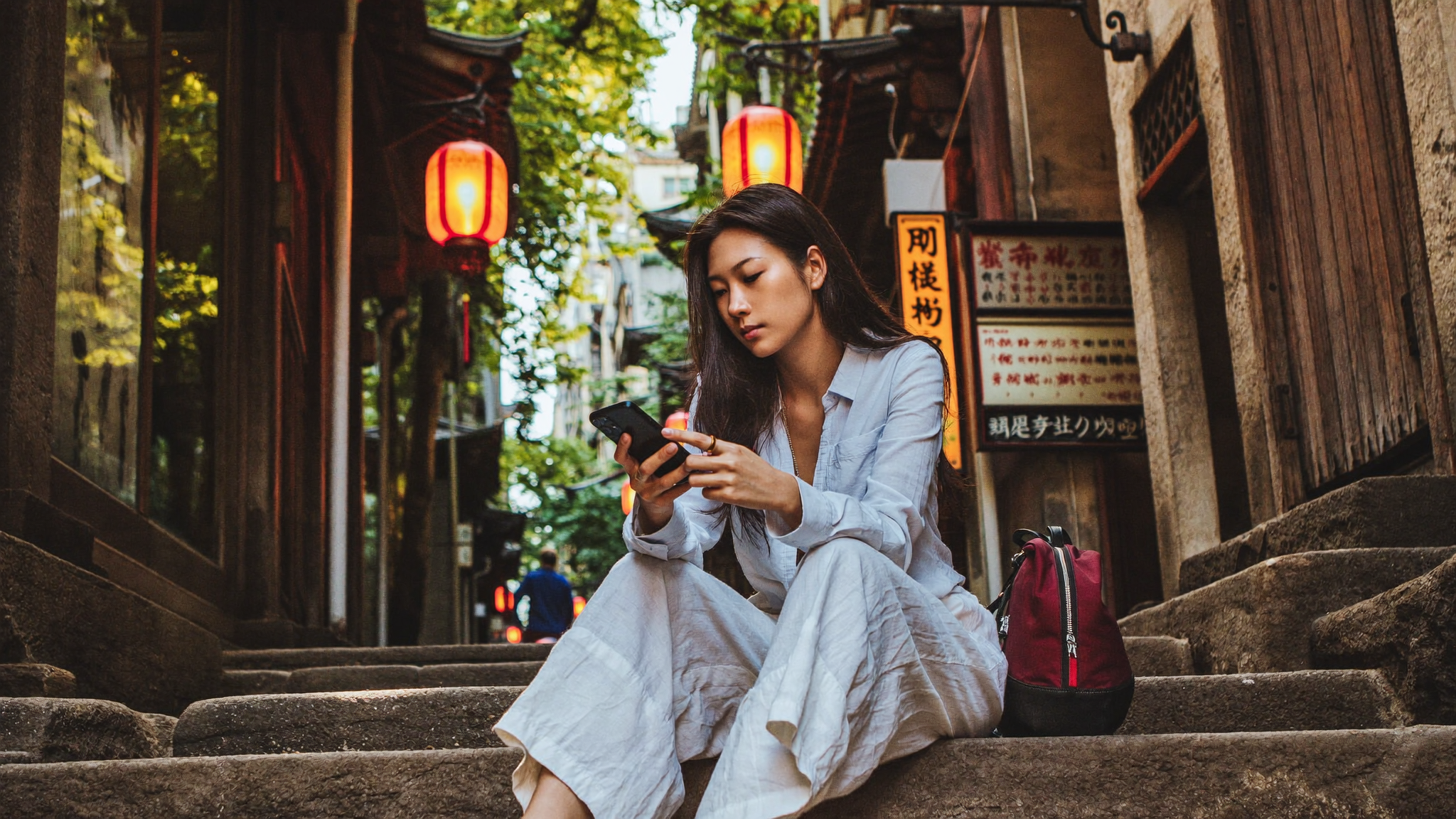 Traveller sitting on stone steps in an old Asian city alley, scrolling on smartphone