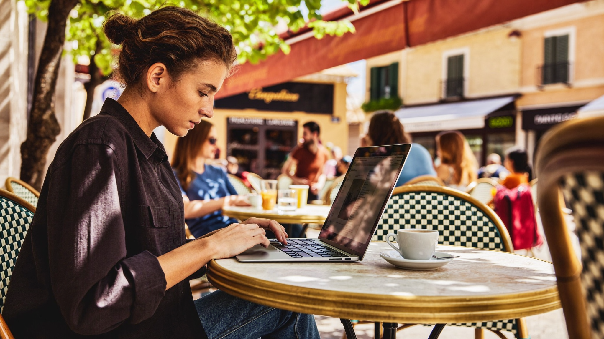 Traveller working on laptop at an outdoor café terrace, bright afternoon light, espresso on the table