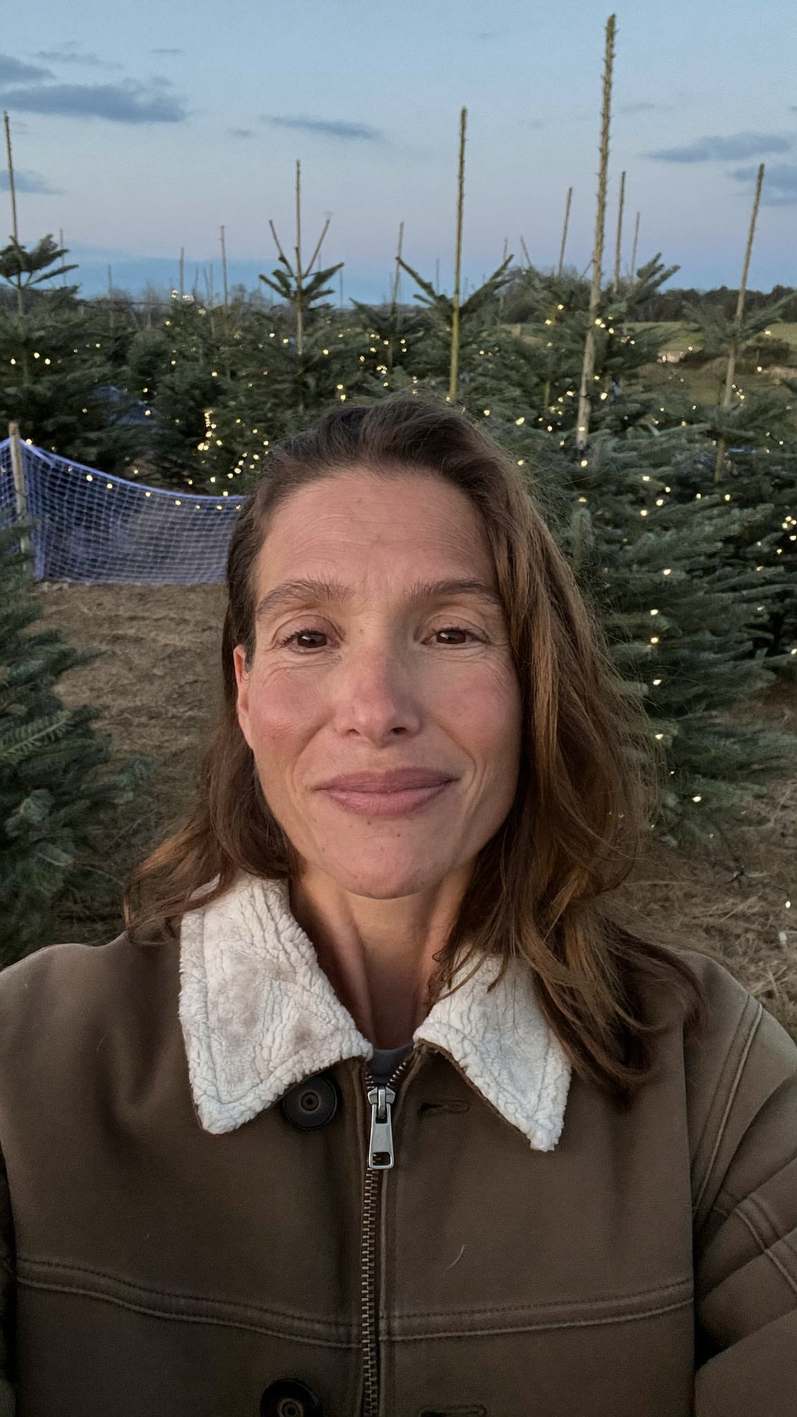 A woman filming herself at a Christmas tree lot at dusk