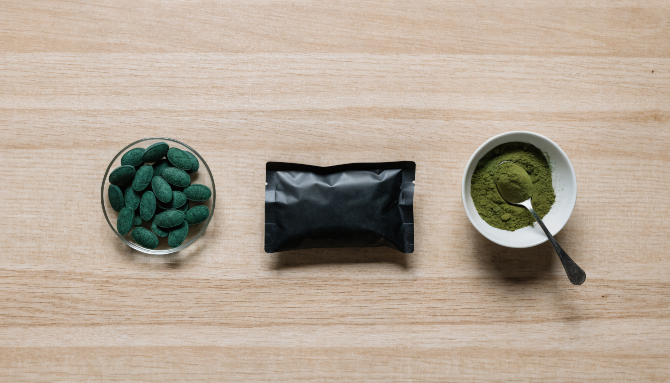 Overhead flat lay comparing a glass dish of green spirulina tablets with a bowl of green spirulina powder beside a matte black supplement pouch
