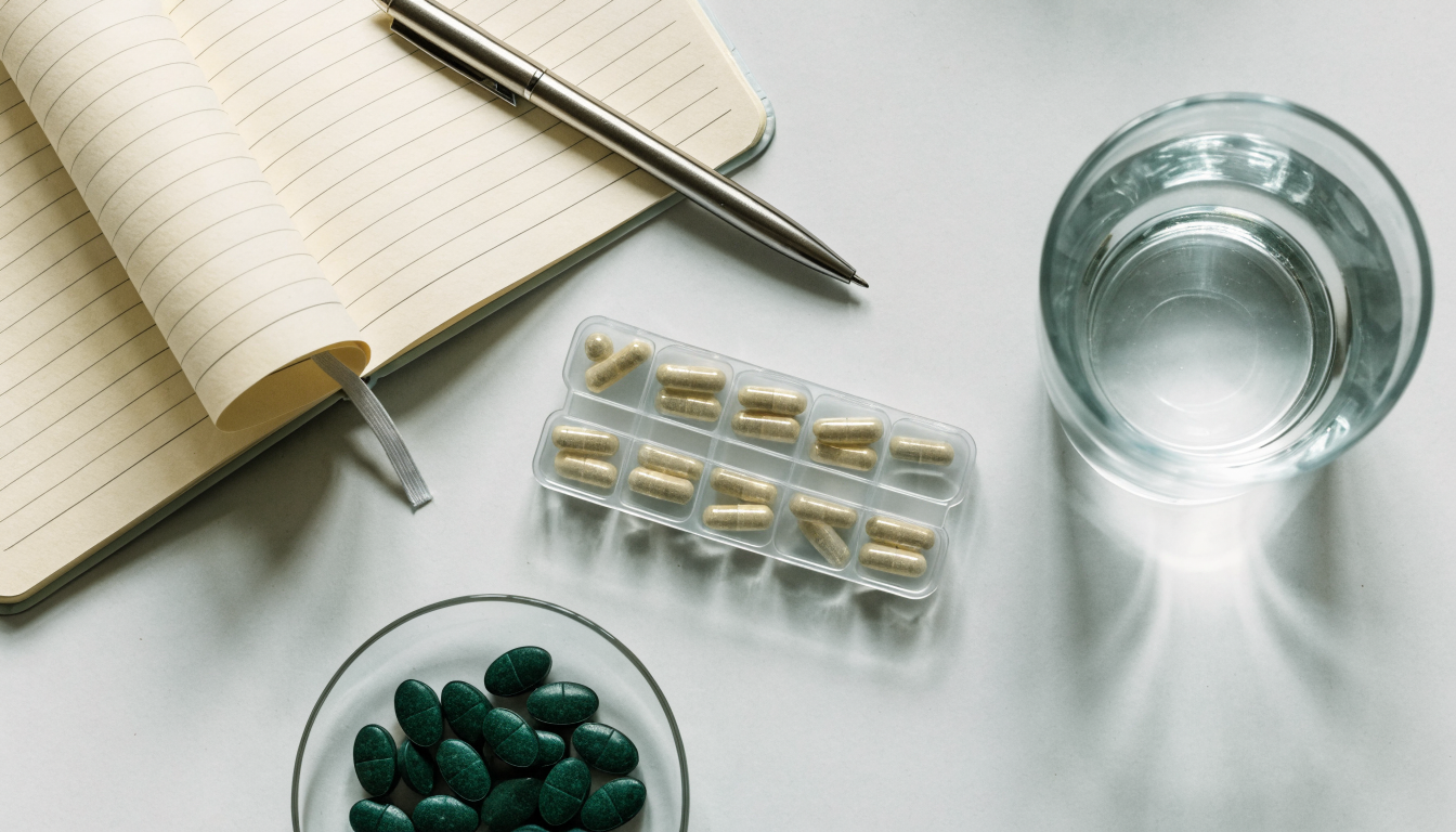 A flat lay of a notebook, weekly pill organizer, and a glass dish of green chlorella tablets on a grey desk