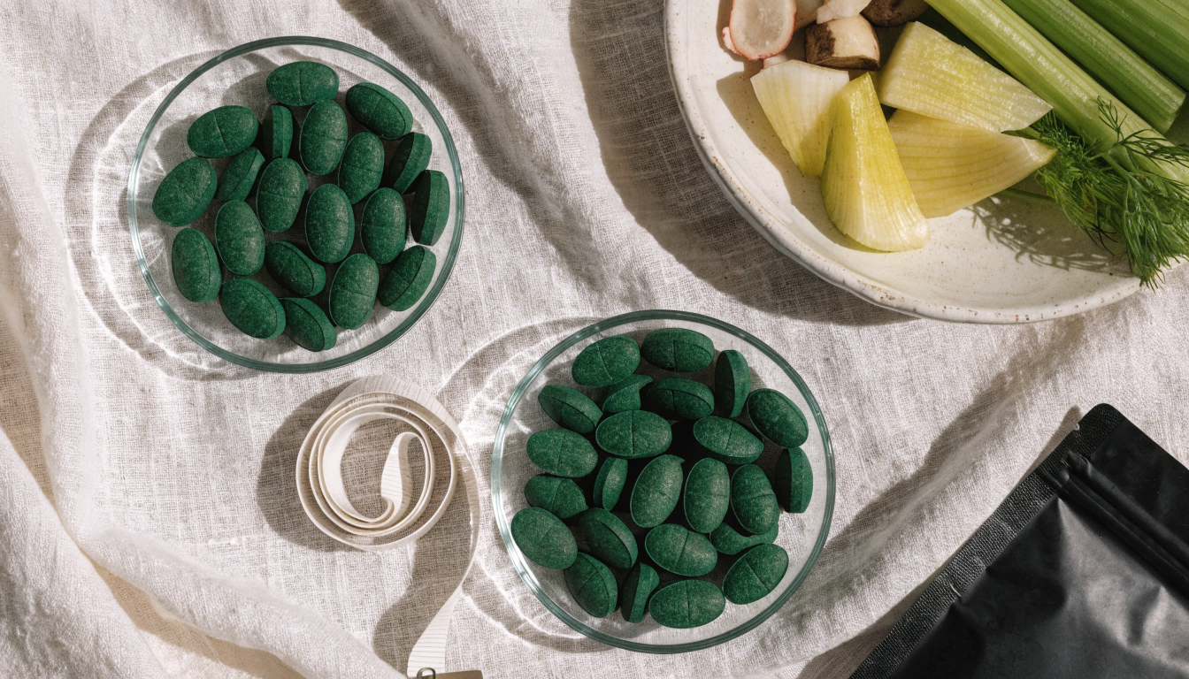 An overhead flat lay on linen with two dishes of green algae tablets, a coiled measuring tape and a bowl of fresh vegetables