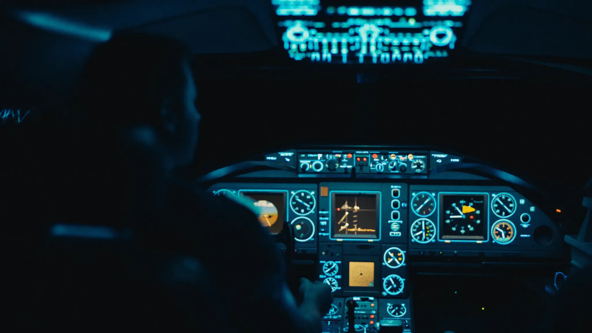 A pilot silhouetted in a dark cockpit, the instrument panel glowing sky blue